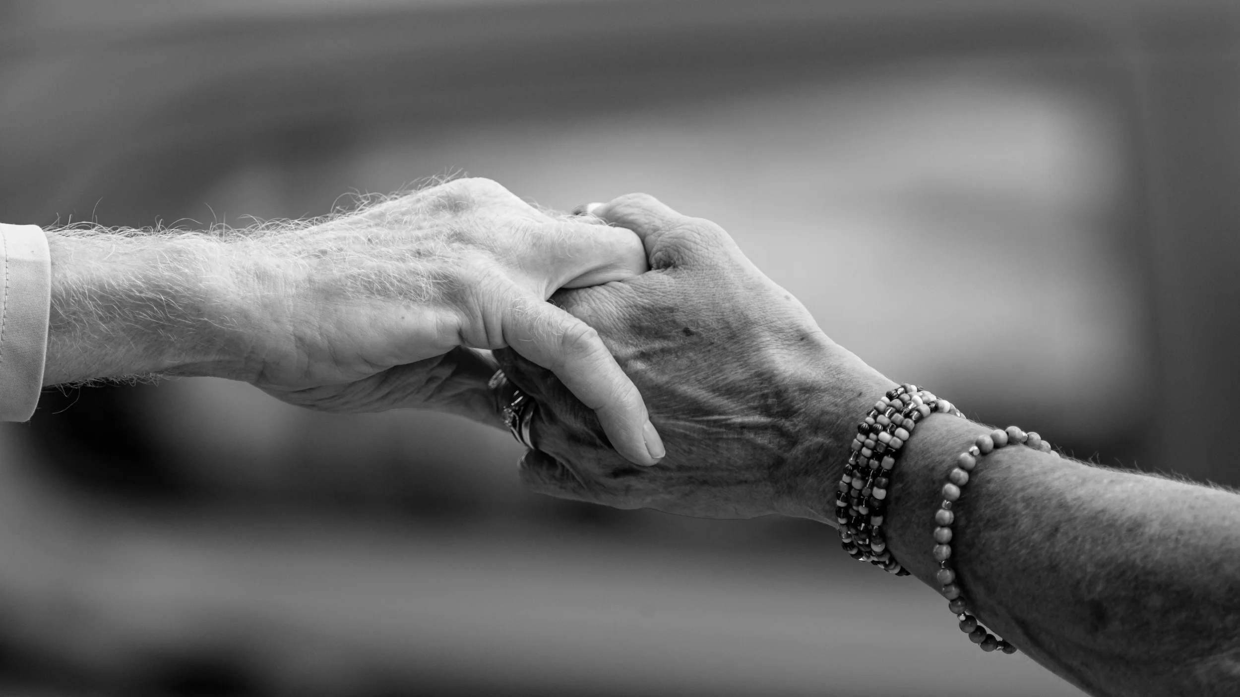Black and white photo of two hands holding each other, one appears older with visible wrinkles and a ring, and the other younger with beaded bracelets.