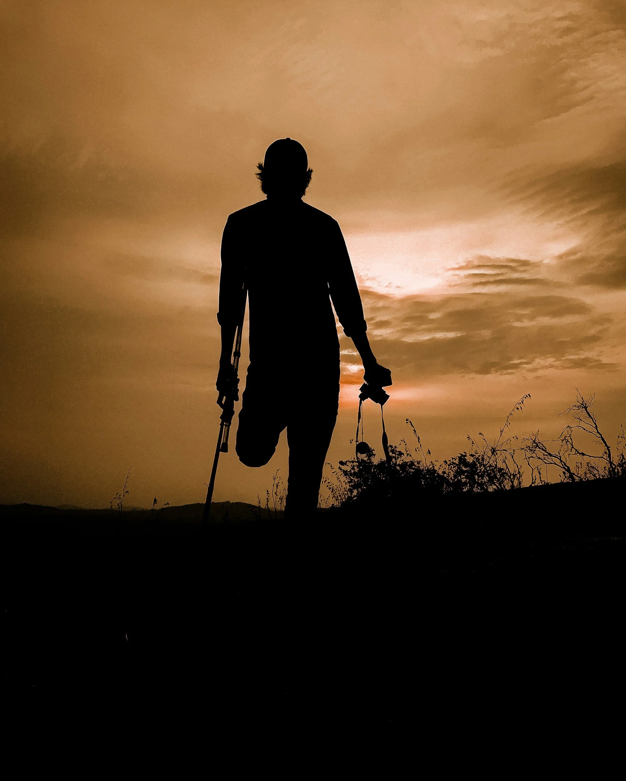 Silhouette of a person holding fishing equipment, standing outdoors during sunset or sunrise with a cloudy sky and trees in the background.