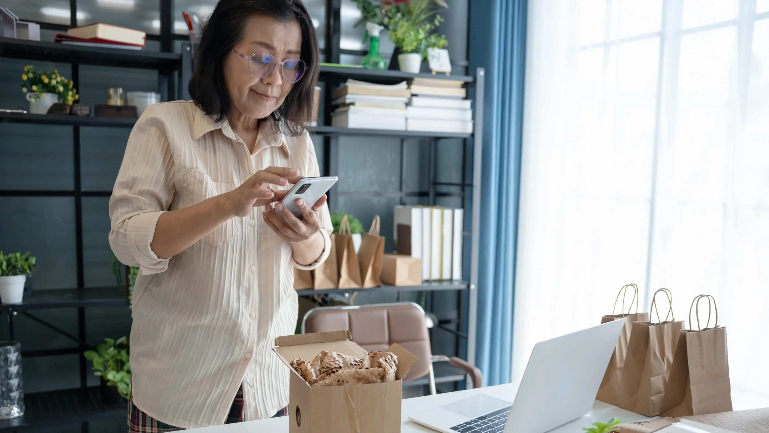 A woman in a beige striped shirt looking at her phone while standing at a table with pizza, paper bags, and a laptop in a bright room with shelves, books, plants, and a window with blue curtains.