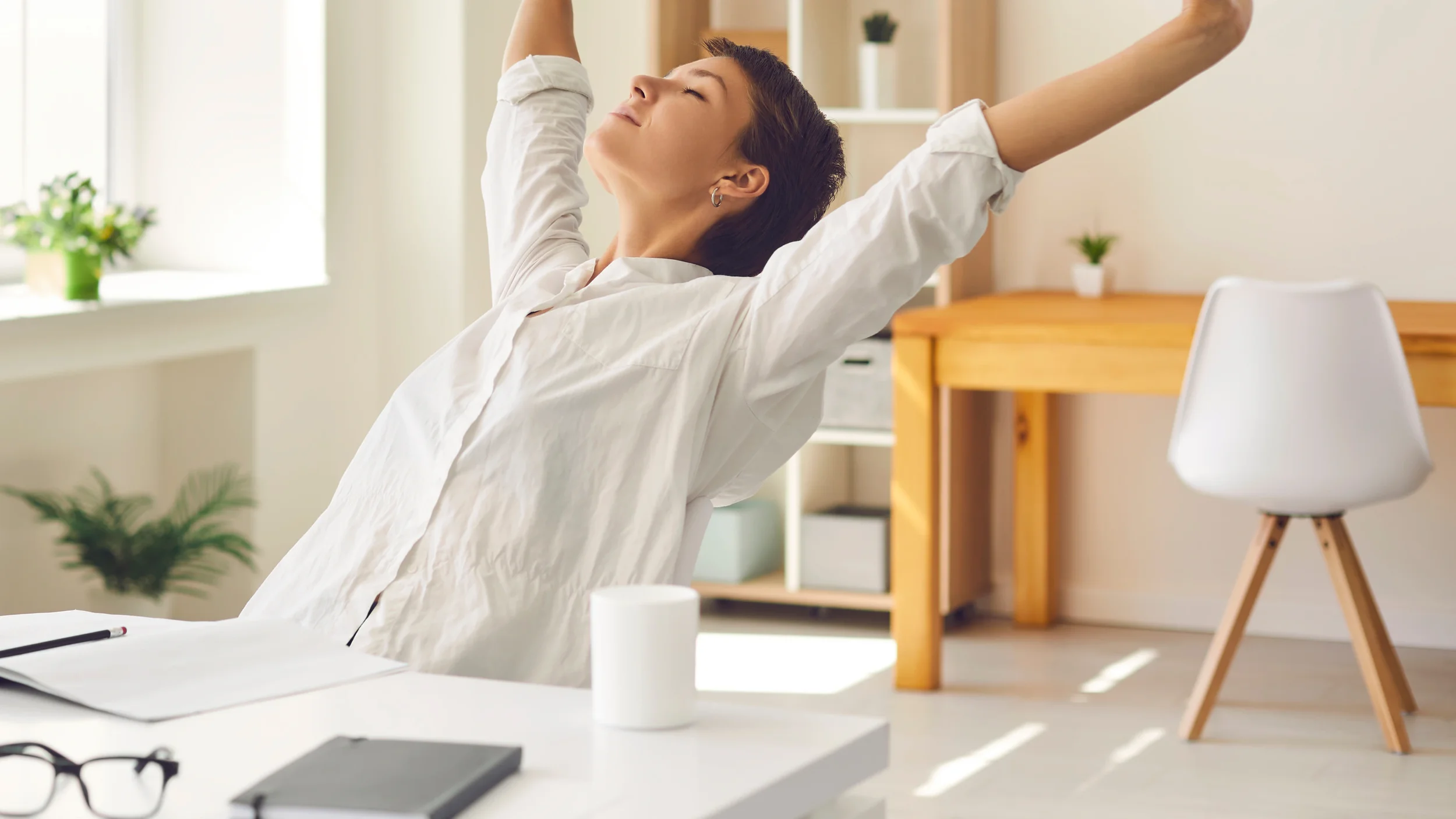 Woman stretching in a bright home office with plants, a wooden desk, and a white chair.