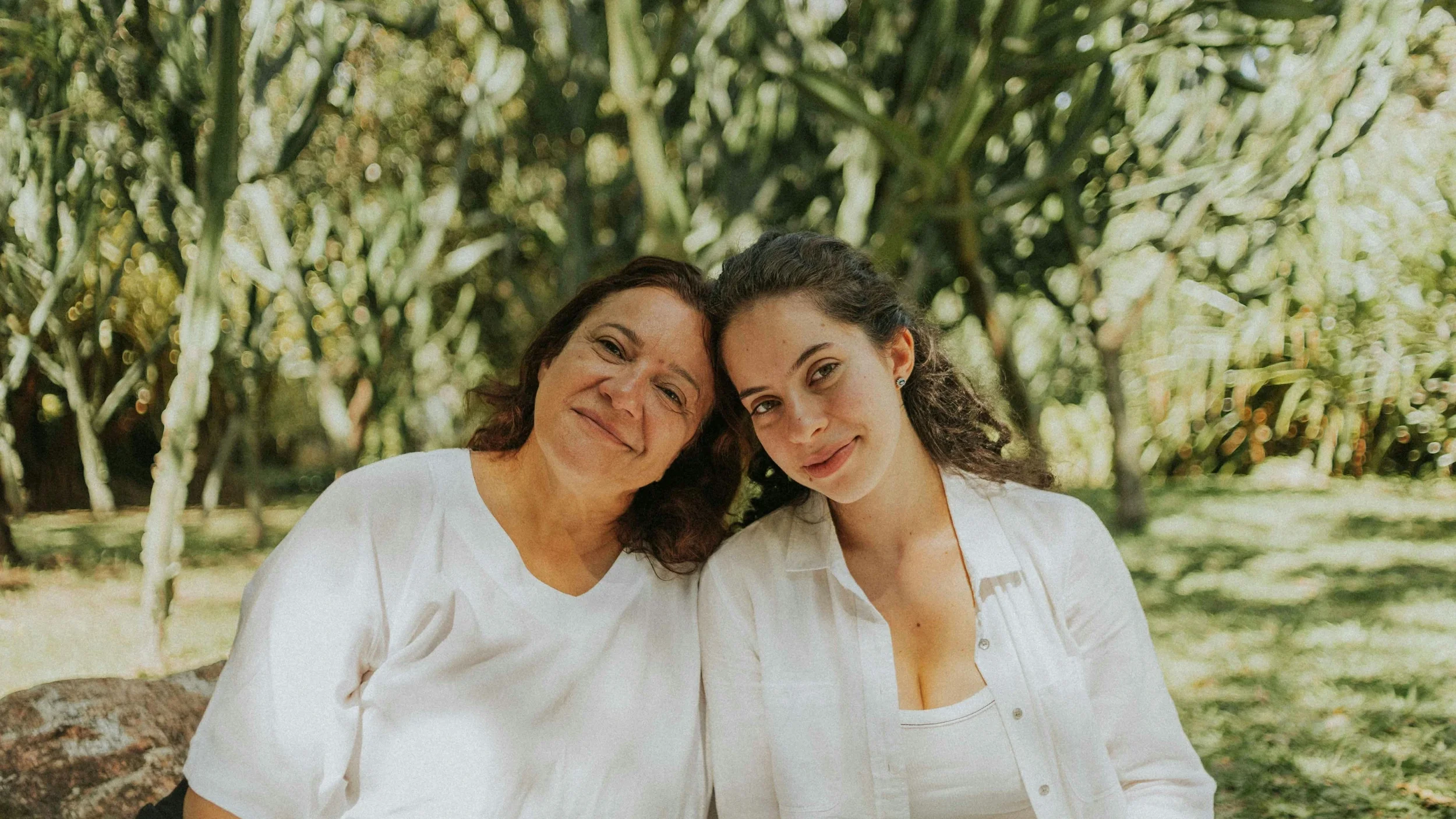Two women, one older and one younger, sitting close together outdoors in a wooded area, smiling at the camera.