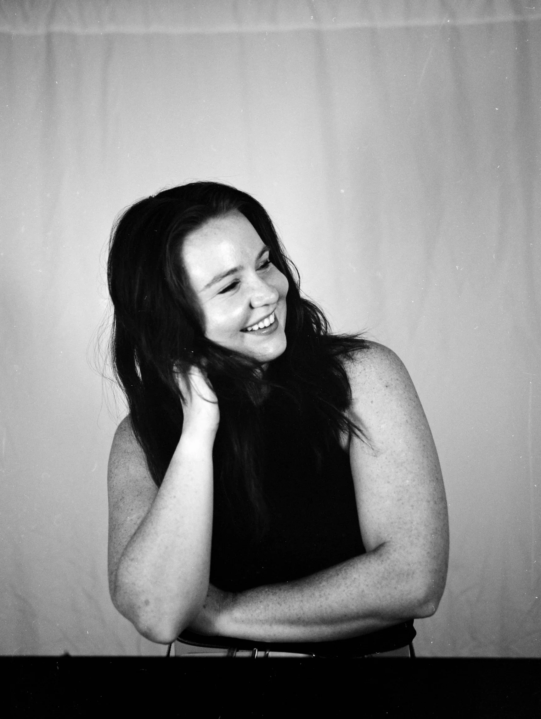 Black and white photo of a woman smiling, with long dark hair, leaning her head on her hand, and looking down.