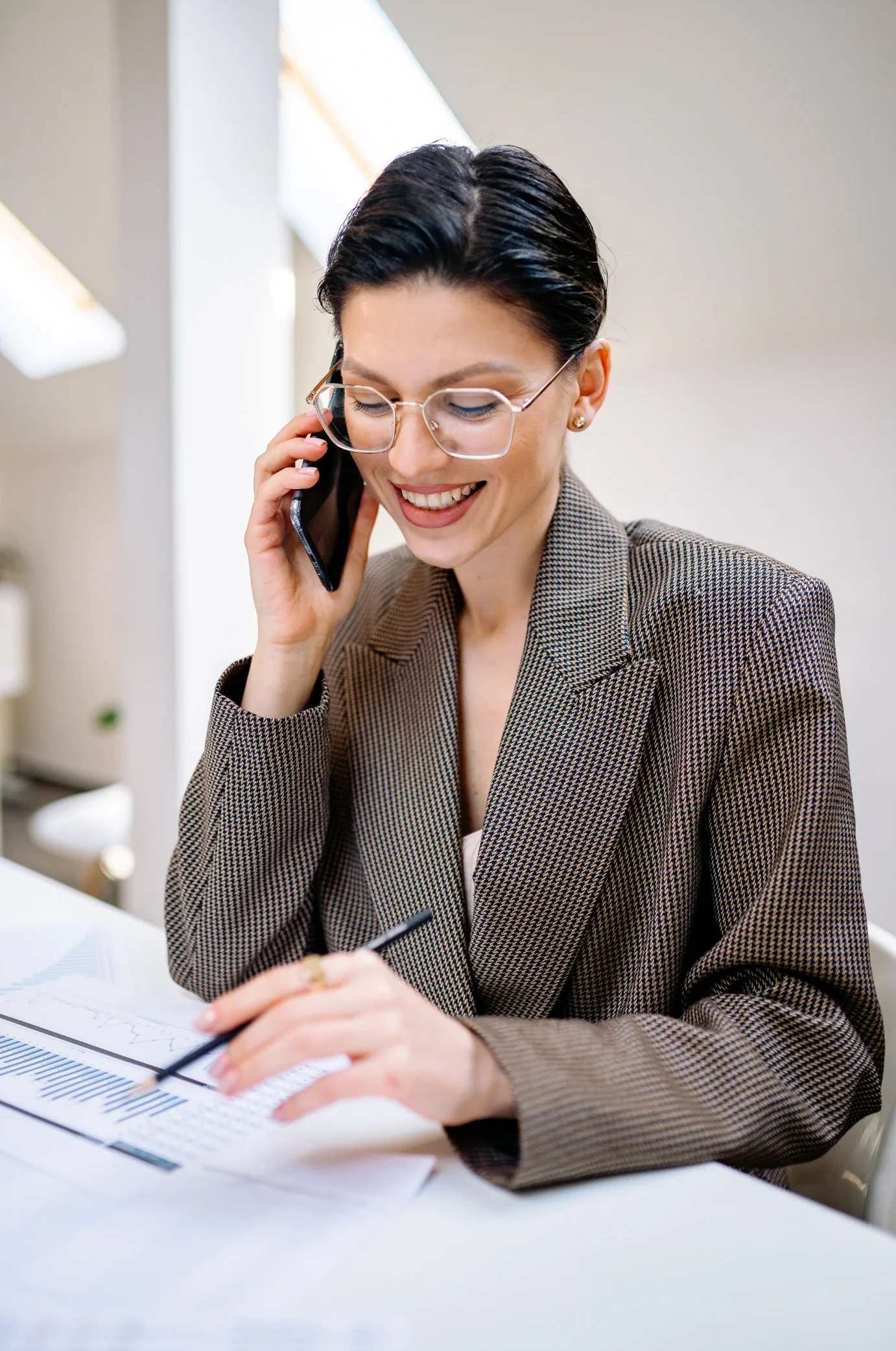 A woman with short dark hair, wearing glasses and a brown checked blazer, is smiling while talking on a mobile phone and writing on papers with a pen at a desk in an office setting.