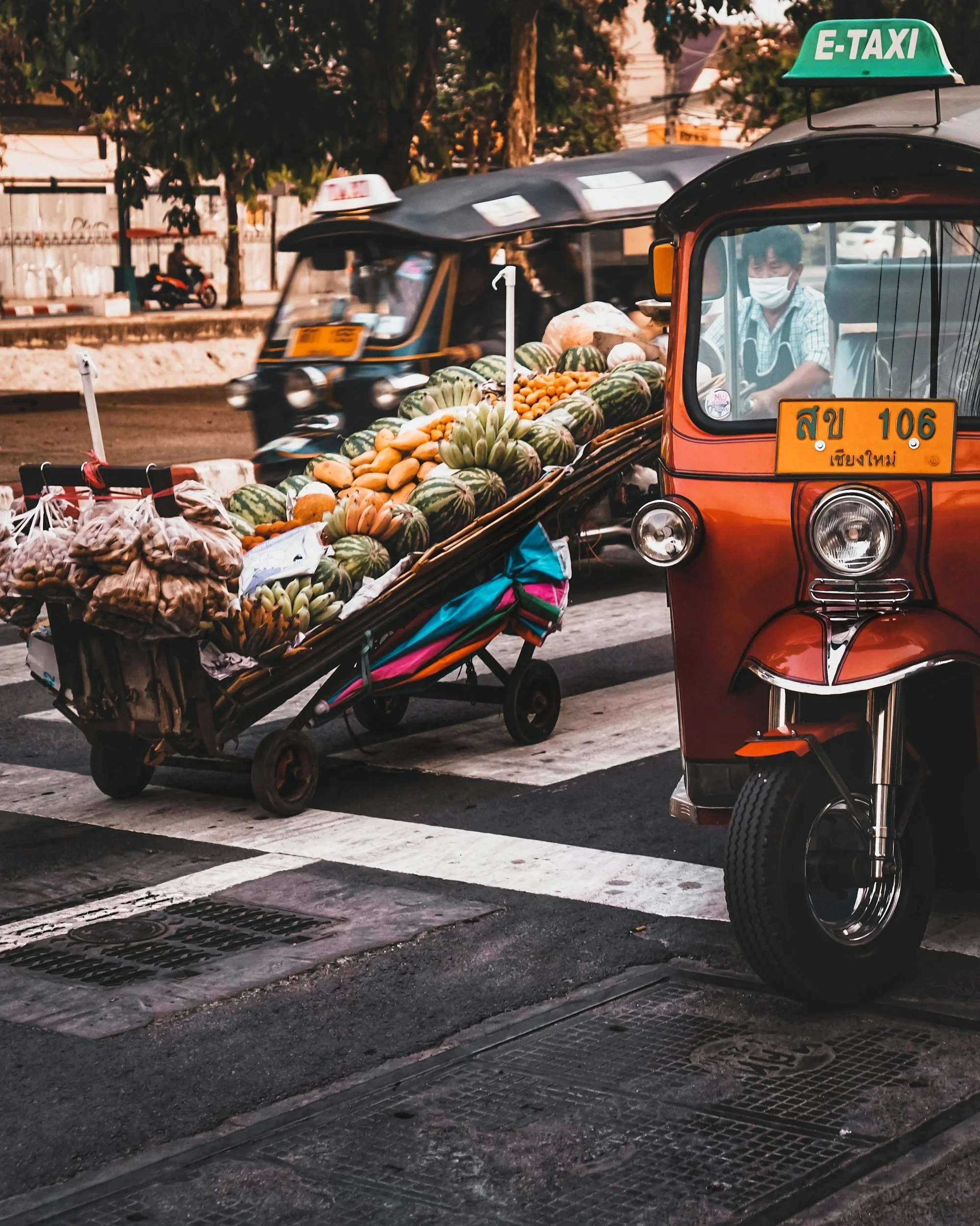 A tuk-tuk vehicle with a sign that says E-TAXI on the roof, parked on a street. The tuk-tuk is carrying a large load of watermelons and other produce. There is a person inside the tuk-tuk wearing a face mask. In the background, another vehicle, possibly a motorized rickshaw or motorcycle, is moving past. Trees and buildings are visible in the background, suggesting an urban setting.