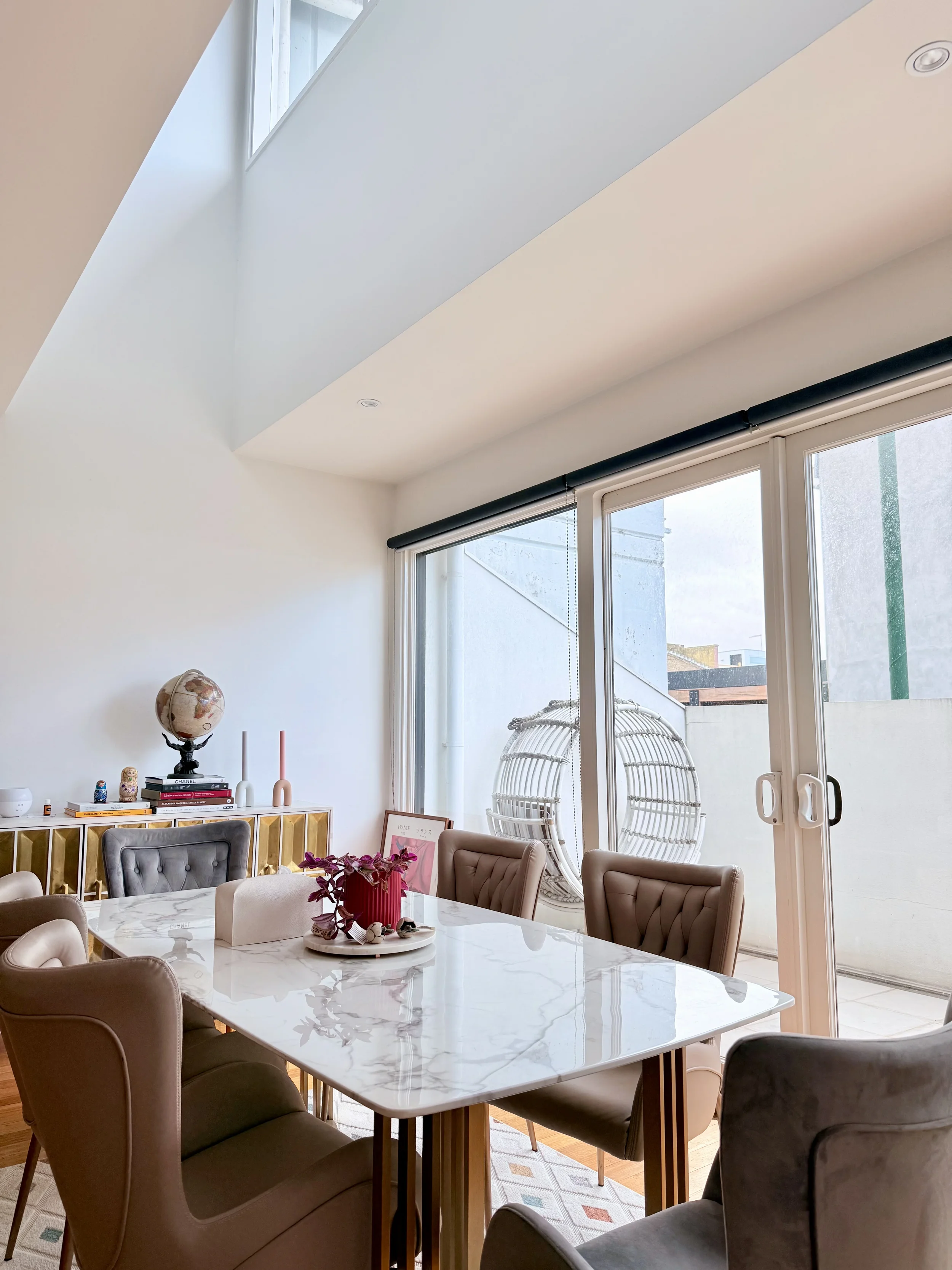 A bright dining room featuring a white marble table surrounded by beige upholstered chairs. A red vase with purple flowers is placed on the table. Large glass sliding doors with black and white roller blinds lead to a balcony with outdoor chair. A white cabinet with books and a globe on top is against the wall. A framed picture is resting on the floor near the cabinet. The ceiling is vaulted with a skylight and recessed lighting.