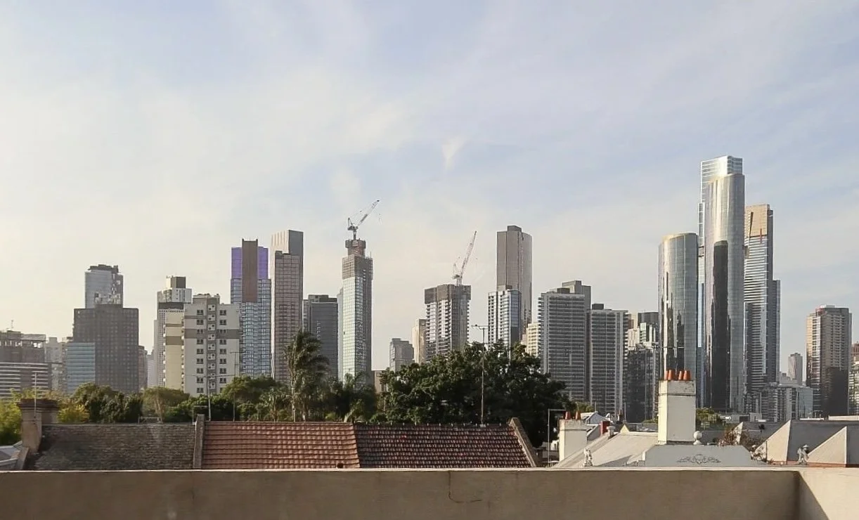City skyline with tall modern skyscrapers under a blue sky, with some construction cranes and rooftops of nearby buildings in the foreground. View from 169 Park Street South Melbourne Ethos Psychology clinic