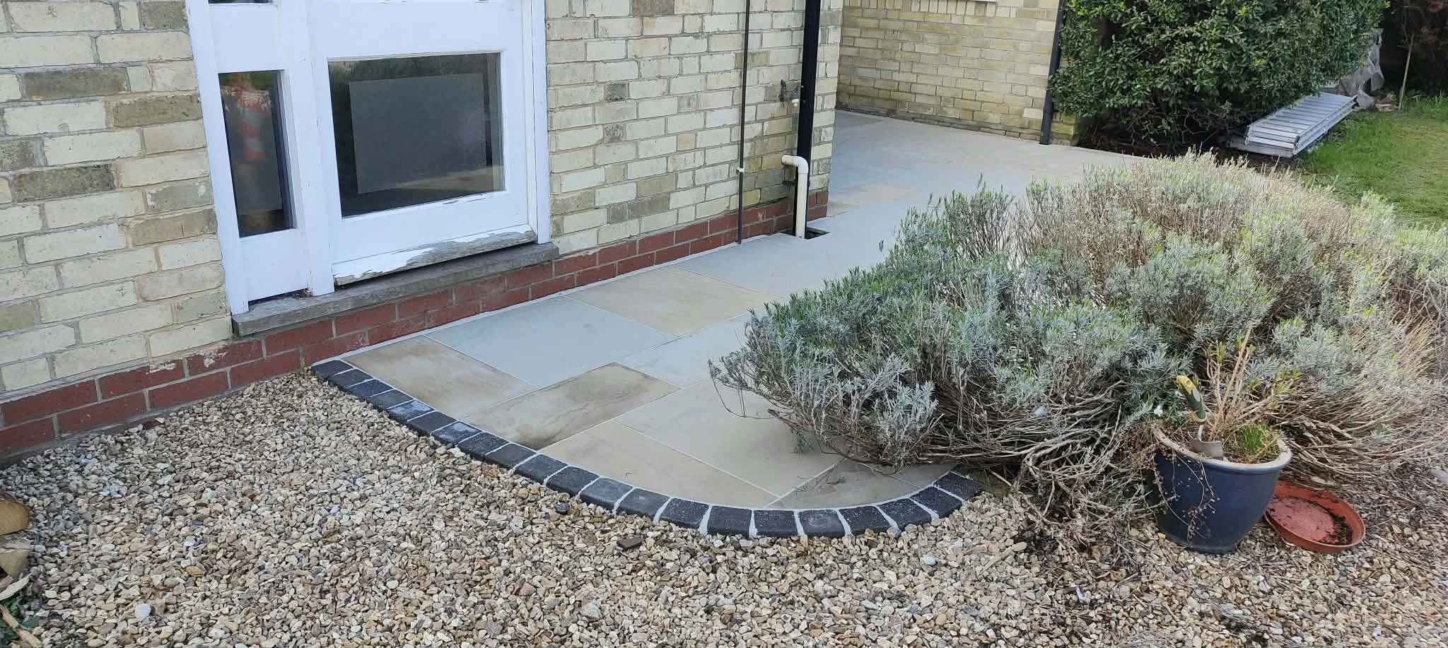 A backyard patio area with a door, potted plants, and a garden bed with dried and green shrubbery, surrounded by small gravel and tile flooring.