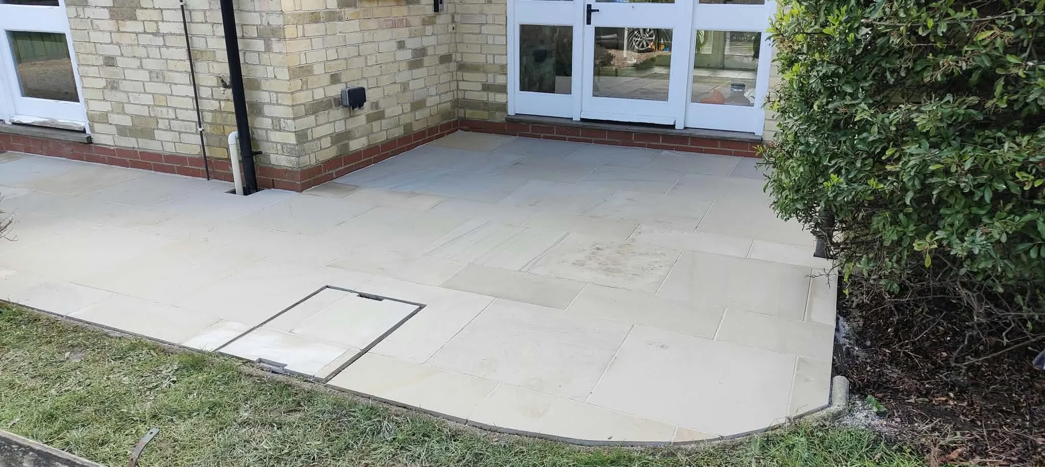 A backyard patio with beige stone tiles, a brick house wall with a glass door, a bush on the right, and a small grassy area in the foreground.