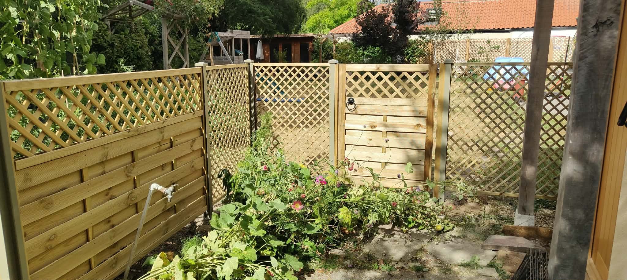 A backyard garden area with a wooden fence and gate, flowering plants, and a hose. There are trees, a shed, and a neighboring house with a tiled roof in the background.