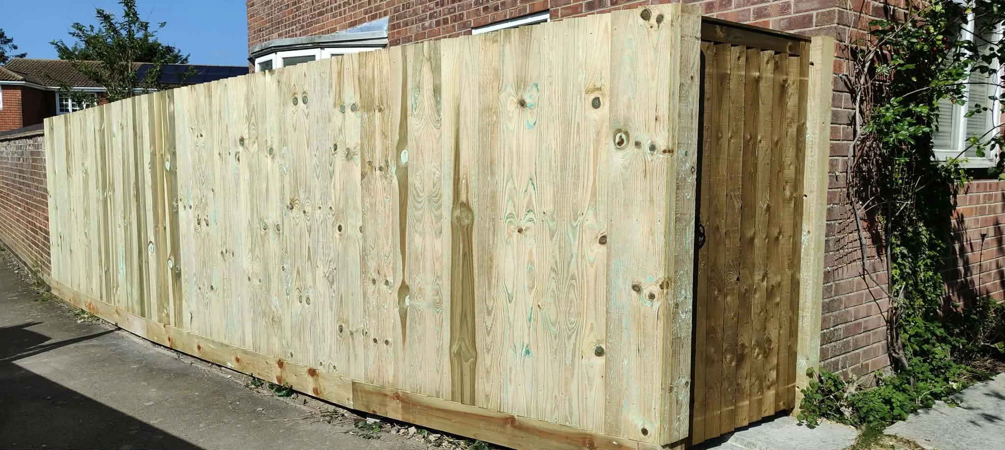 New wooden privacy fence being installed along a brick house with a side door and next to a vine-covered wall.