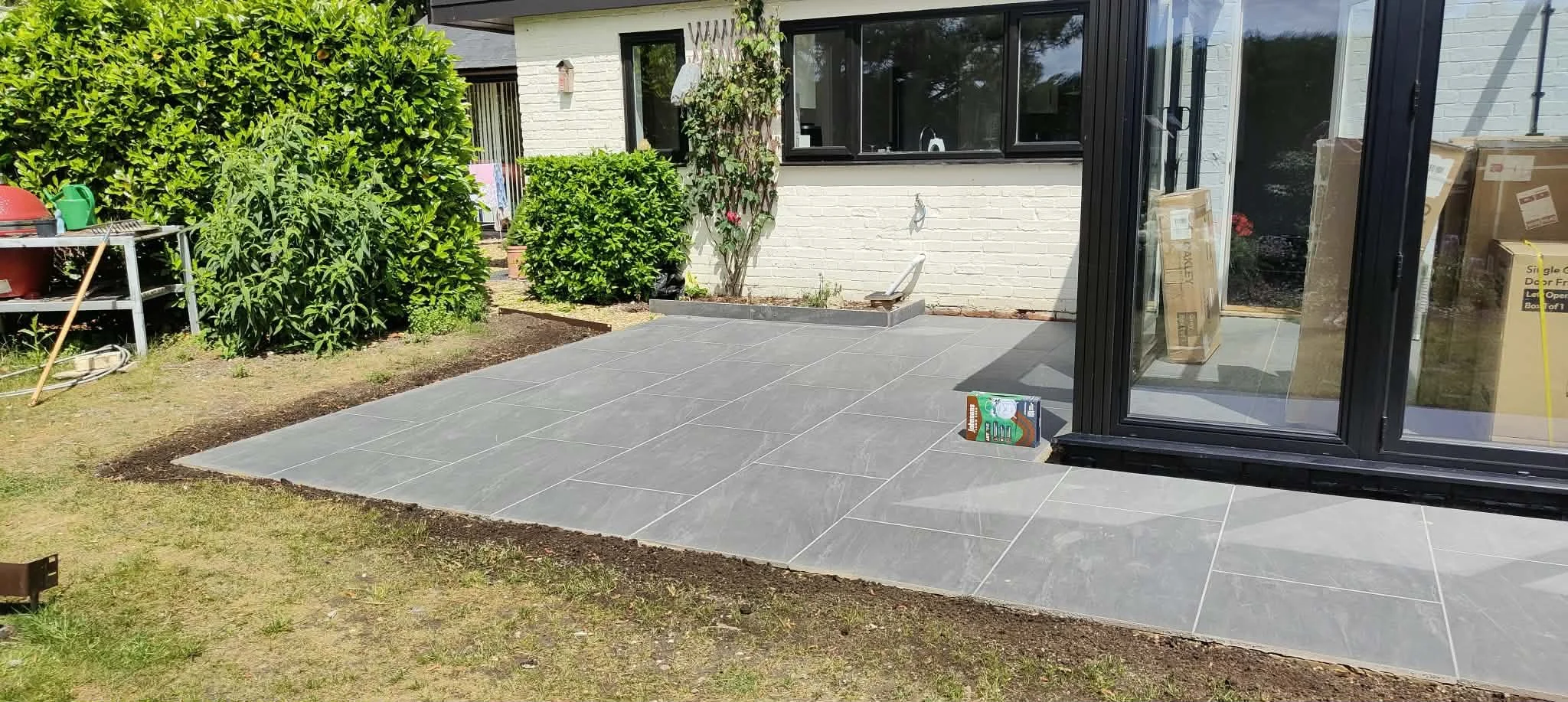 Backyard patio under construction with gray tiled flooring, adjacent to a white brick house, and bordered by green bushes and a grassy yard. There are construction materials and tools nearby.