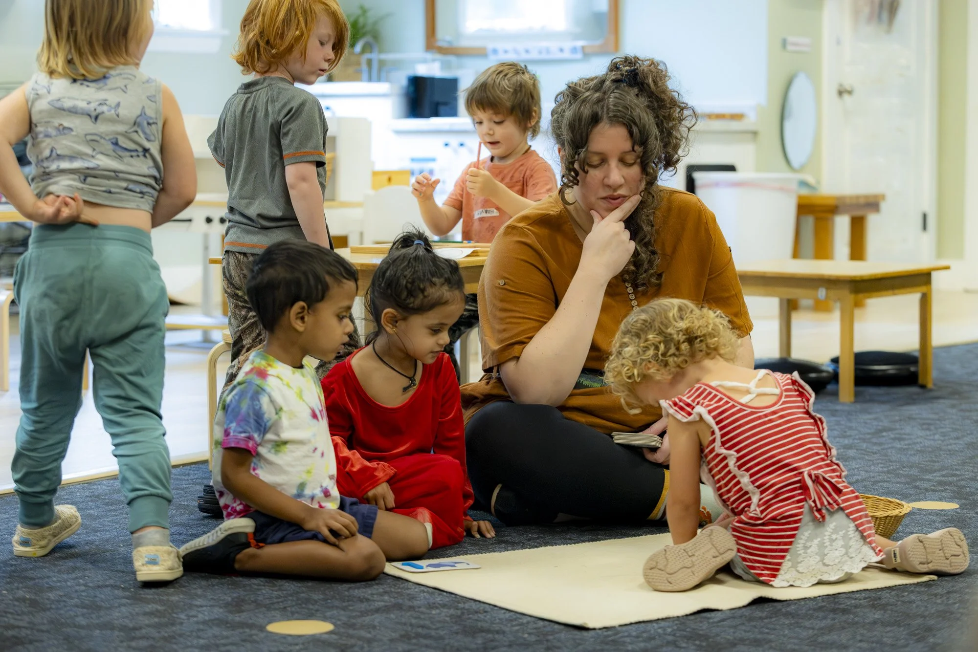 A teacher or caregiver sitting on the floor with young children in a classroom, some seated on a rug, engaging with a child in a red and white striped dress who is kneeling and looking at something on the rug.