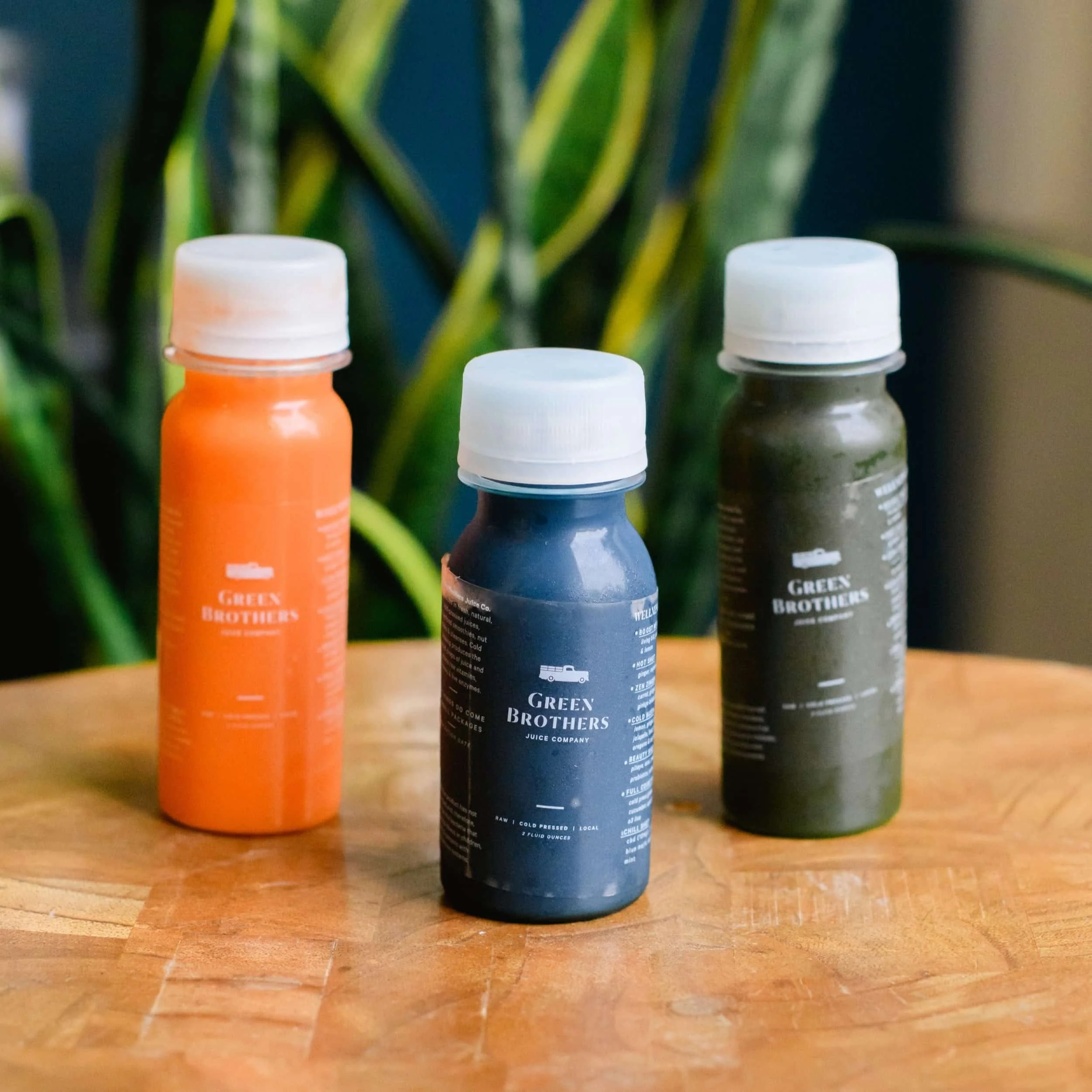 Three bottles of juice labeled 'Green Brothers' on a wooden table with a green plant in the background.