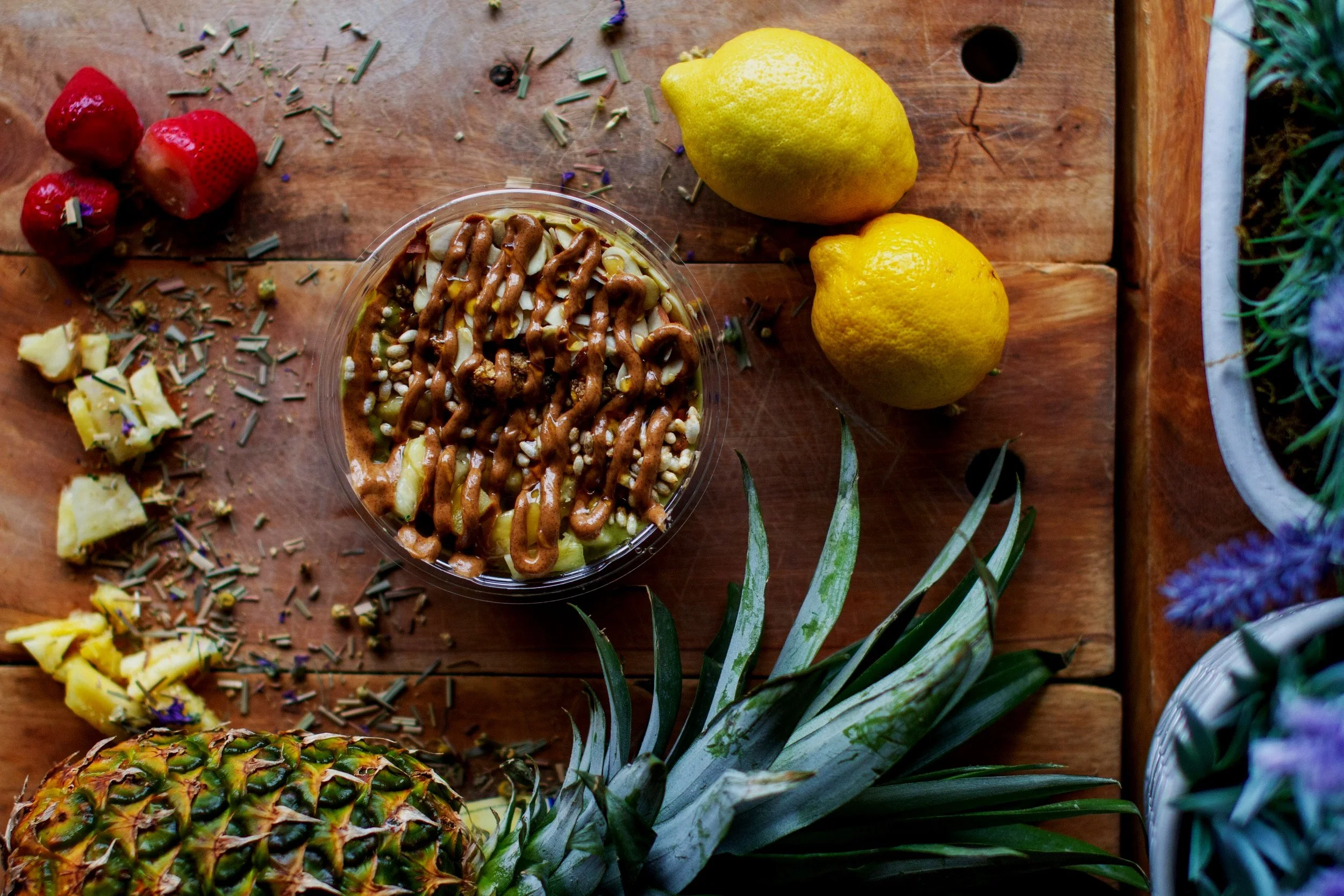 A wooden table with pineapple, two lemons, strawberries, pineapple chunks, herbs and a pineapple-shaped plant in the background.
