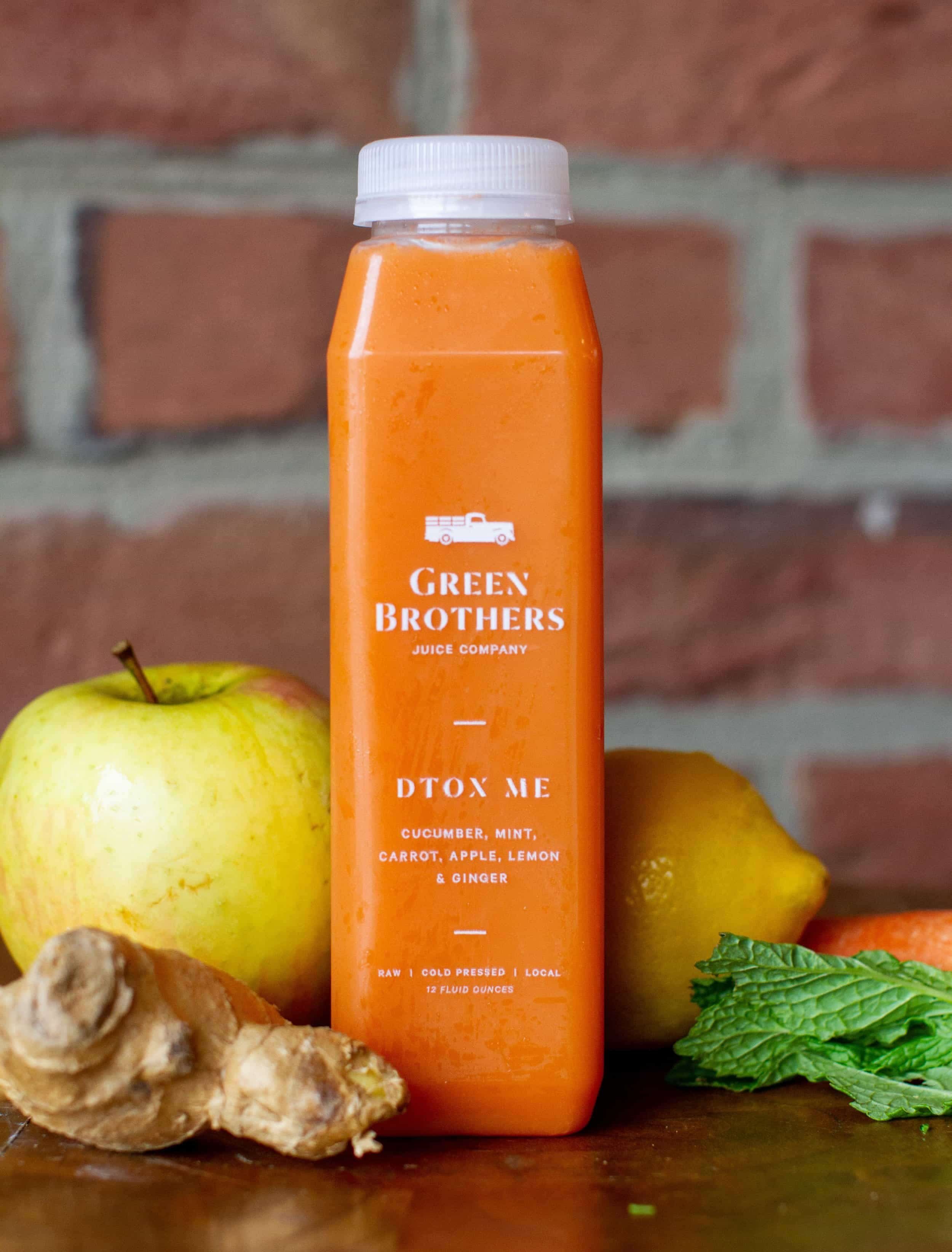 Orange juice bottle labeled Green Brothers Detox Me, a detox juice, sits among apple, lemon, ginger, carrot & mint by a brick wall.