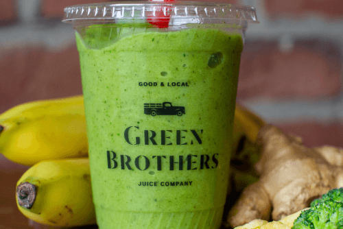 A green smoothie in a plastic cup with a red straw, surrounded by bananas and ginger, labeled Green Brothers Juice Company.
