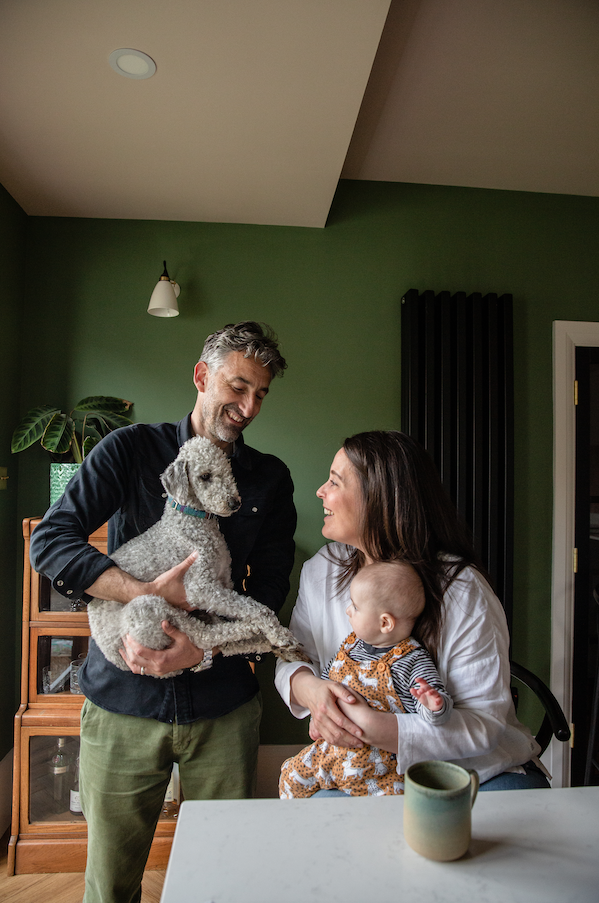 family sat in the kitchen. Dad is holding a Bedlington Terrier while mum is holding the baby