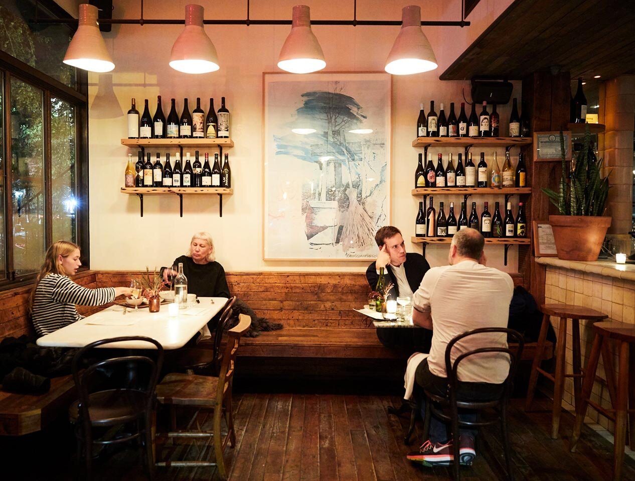 Two tables enjoy Rhodora's evening atmosphere. Windows to the left overlook a Brooklyn street, and behind the patrons are wine bottles along the wall. 