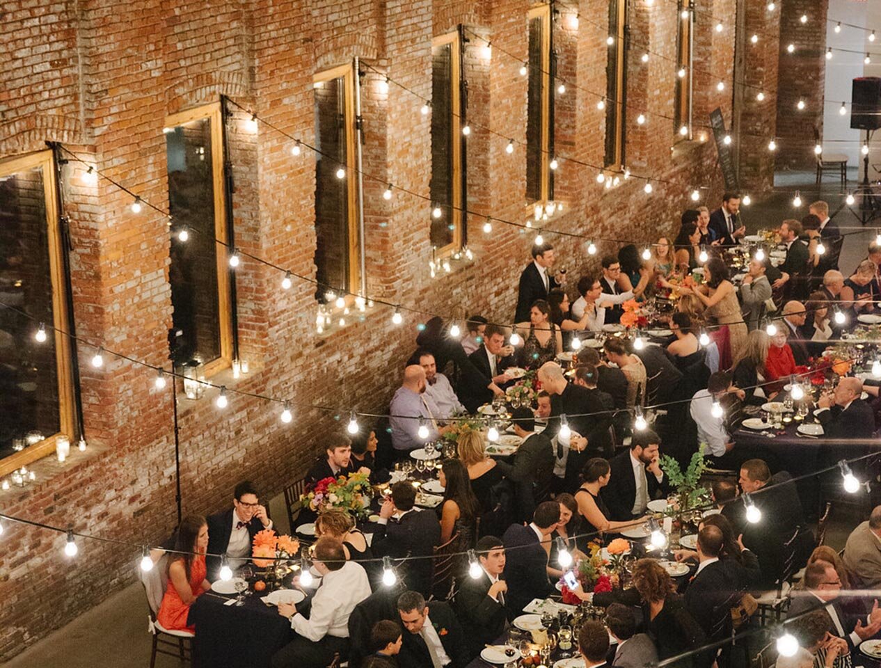 Bird's eye view of guests at Pioneer Works sharing food at long tables underneath strings of Edison bulbs. 