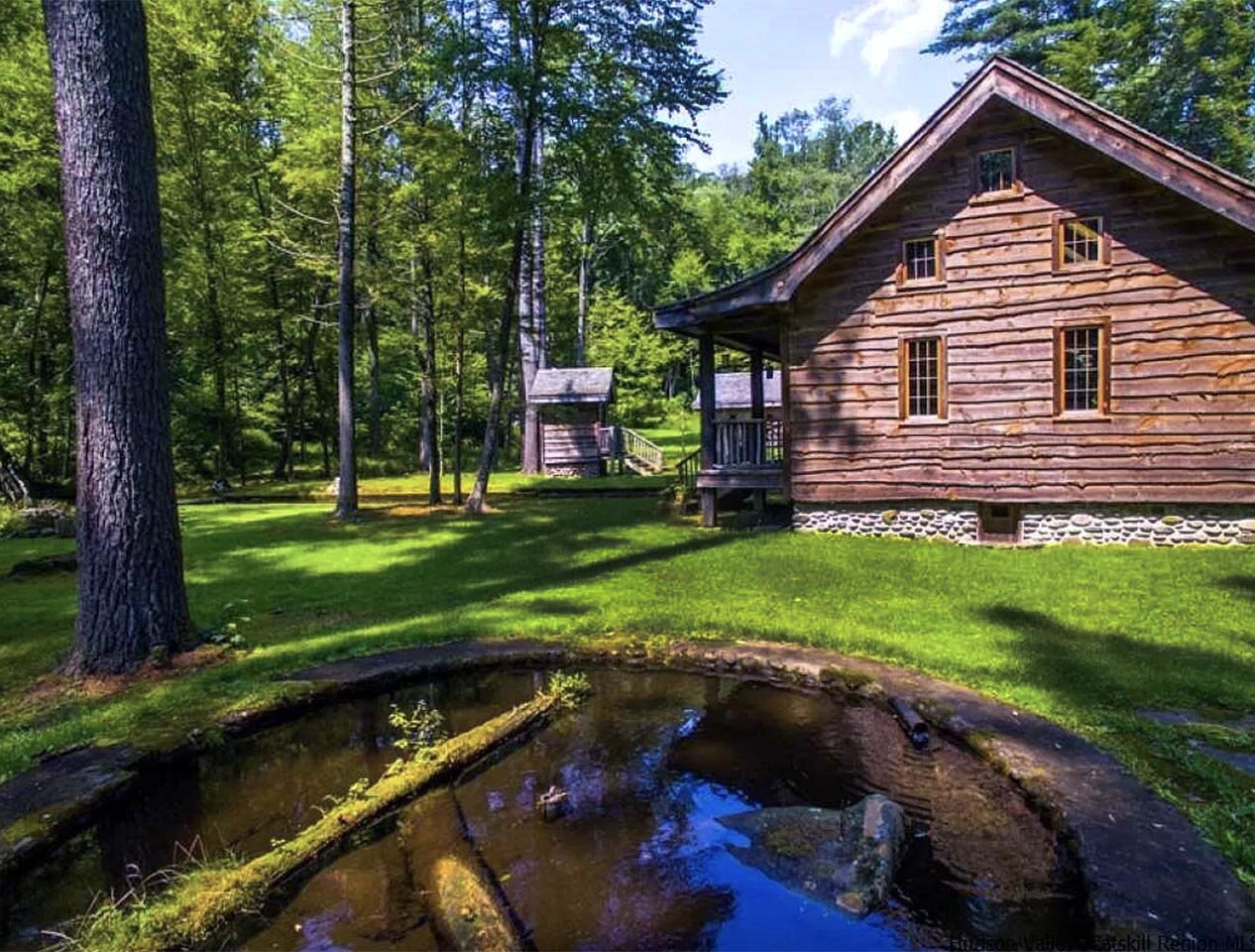 A rustic wooden historic cabin with a stone foundation at the Jenny Brook River School, with a pond in the foreground.
