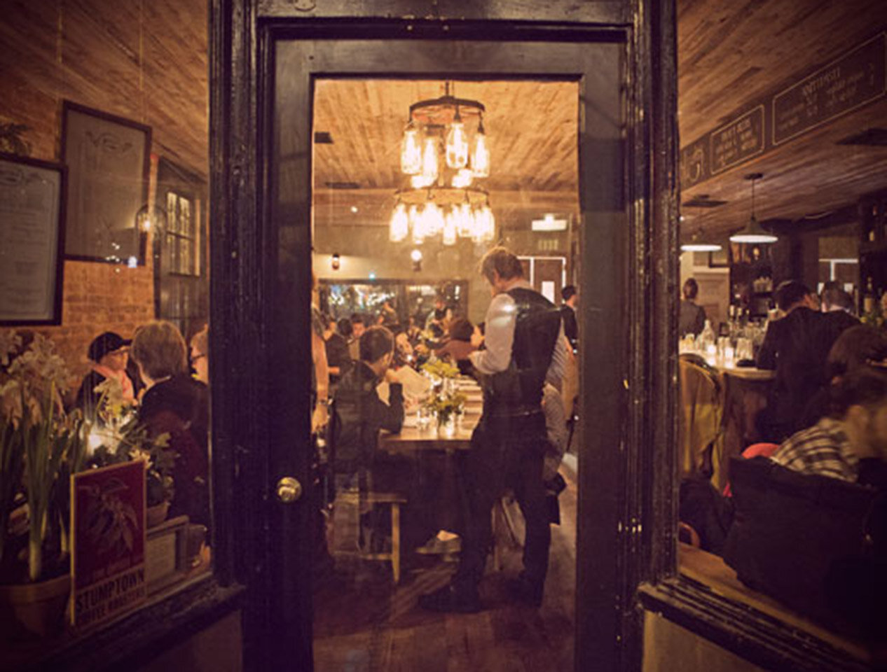 View through the front windows of a busy Brooklyn restaurant called Rucola. It's evening, and the lights inside are bright. 