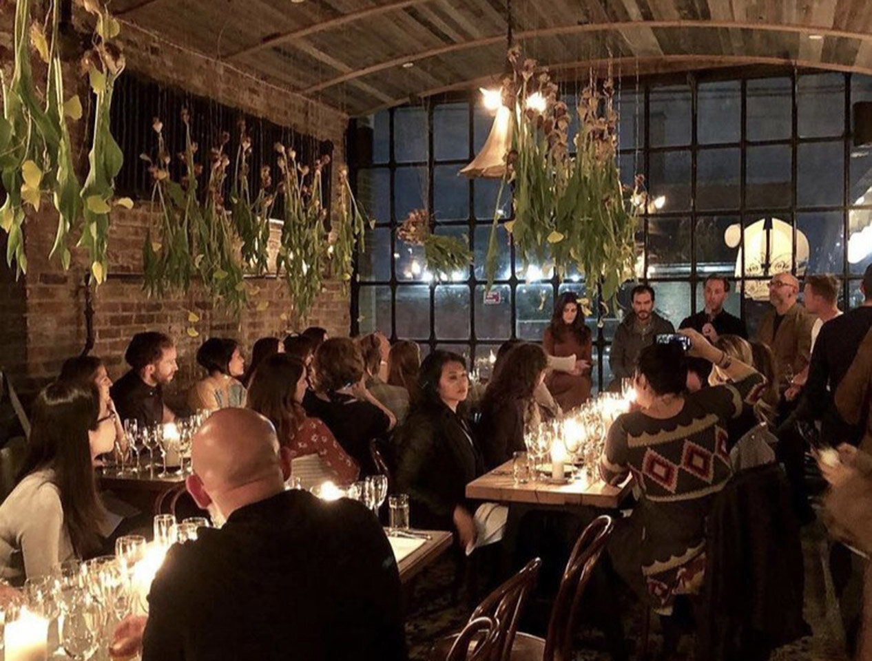 Tables of patrons listen to a speech from Chef Doug McMaster at Silo NYC, with flowers handing above from the ceiling
