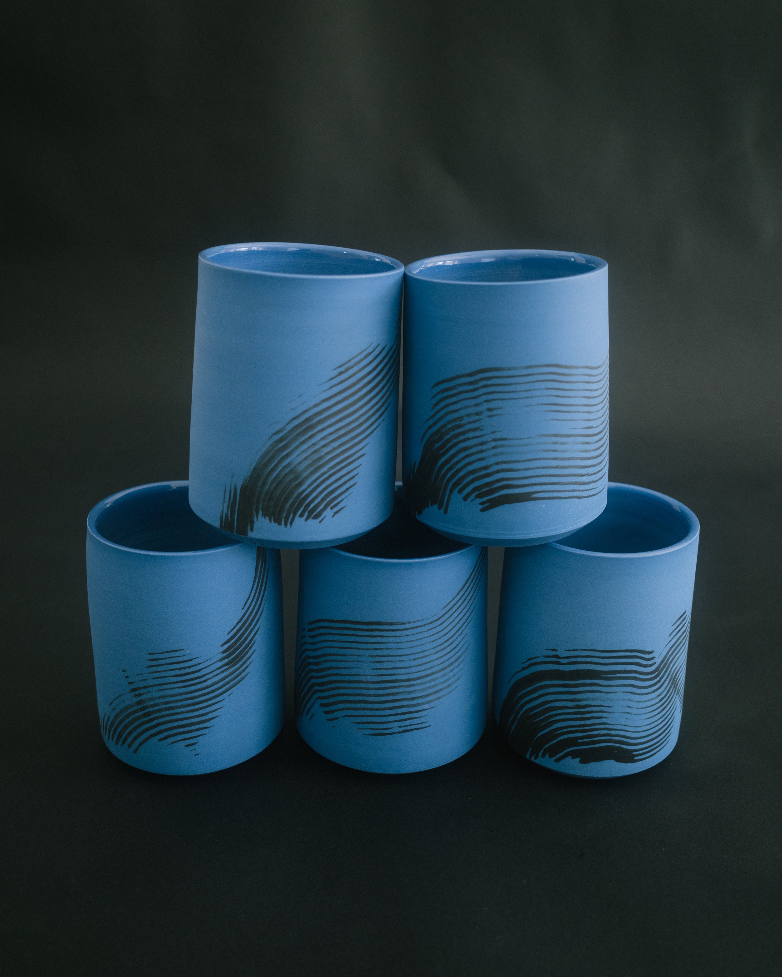 5 blue ceramic tumblers stacked in a pyramid formation. Two on top and two on the bottom on a black seamless background. They each have a black brush stroke on the exterior.
