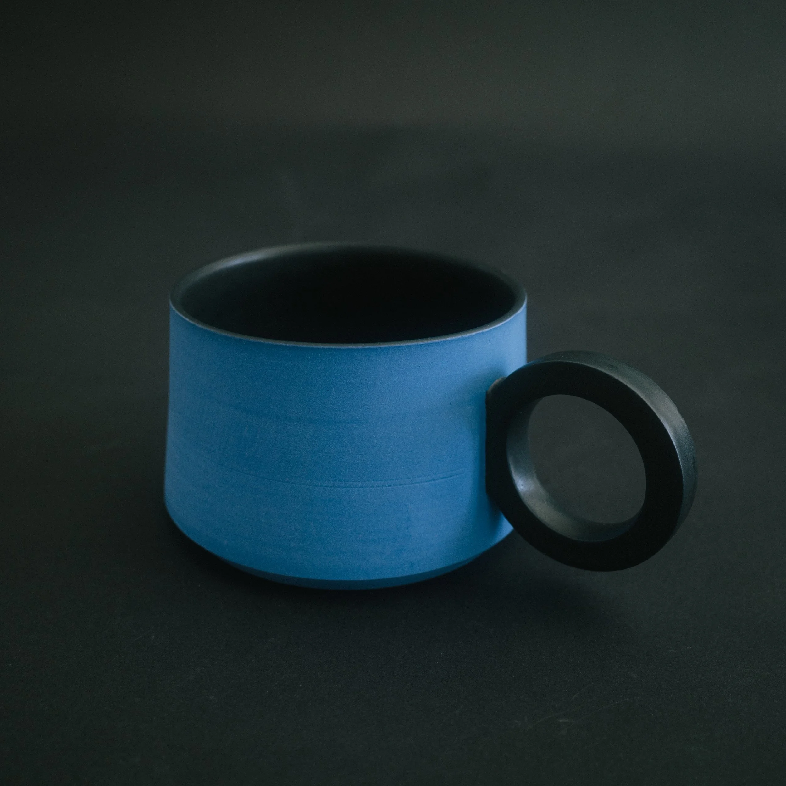 A single blue ceramic mug with a black handle and black interior on black a black seamless background.