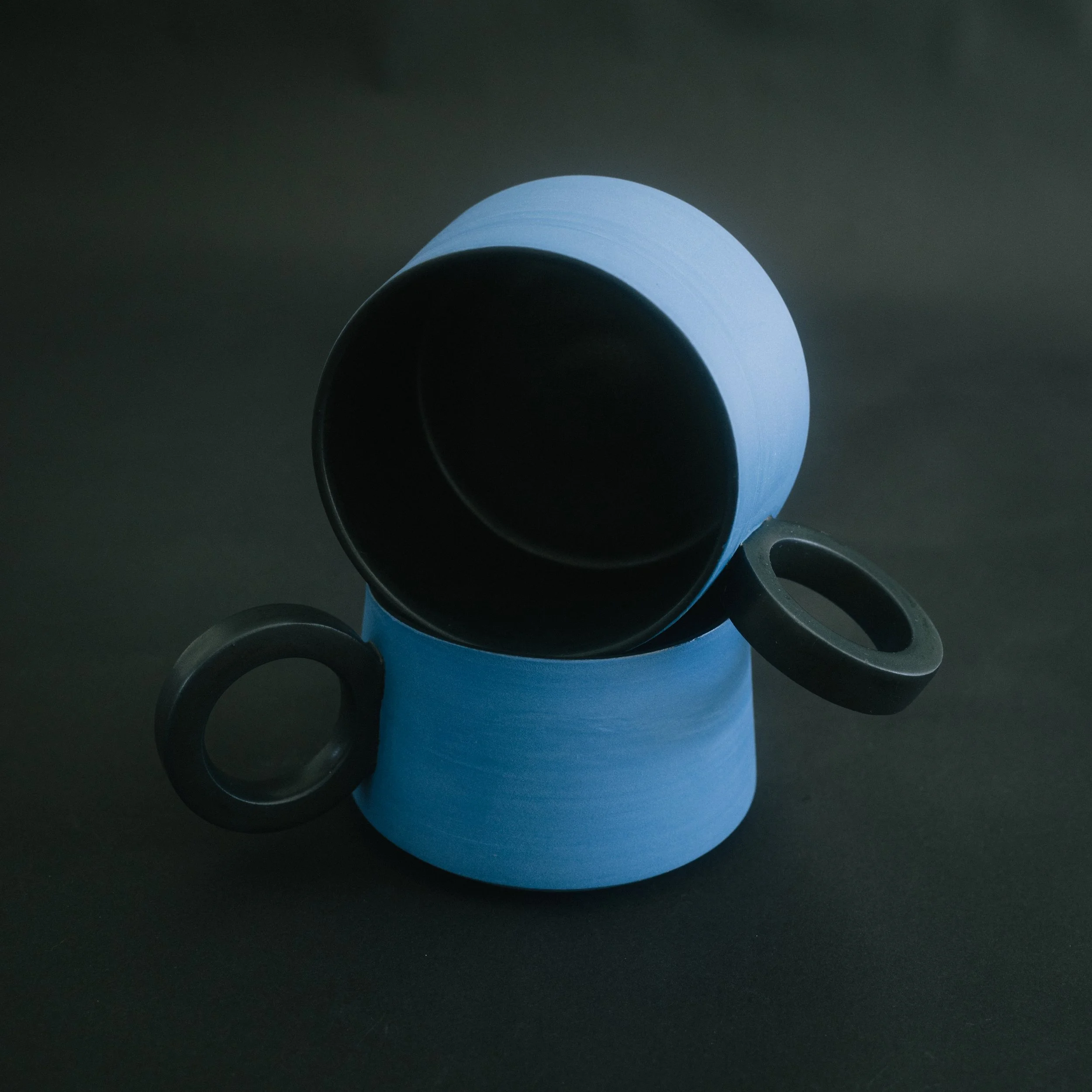Two blue ceramic mugs with black handles and black interiors on black a black seamless background.