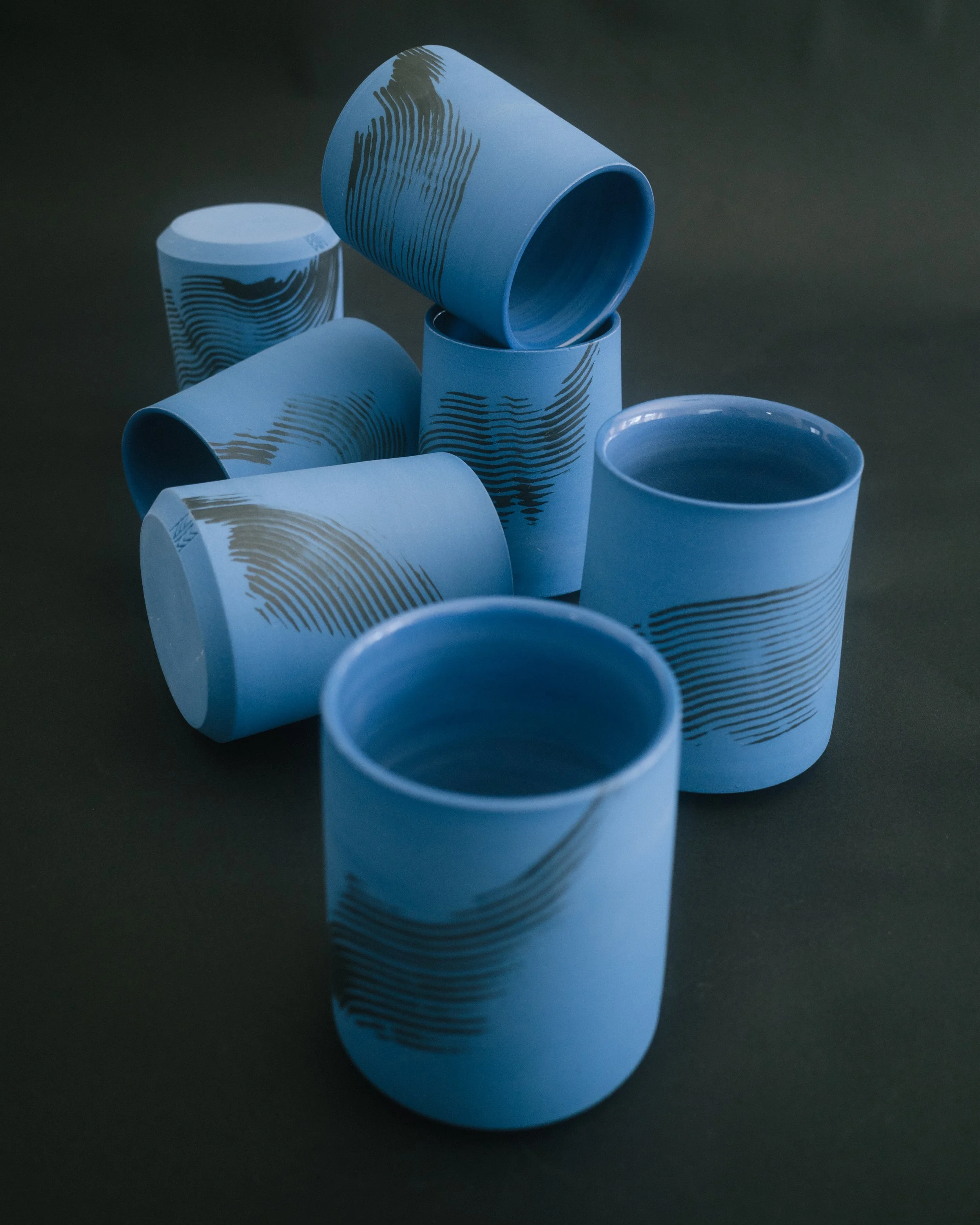 A group of blue ceramic tumblers with black brush strokes on a black seamless background.