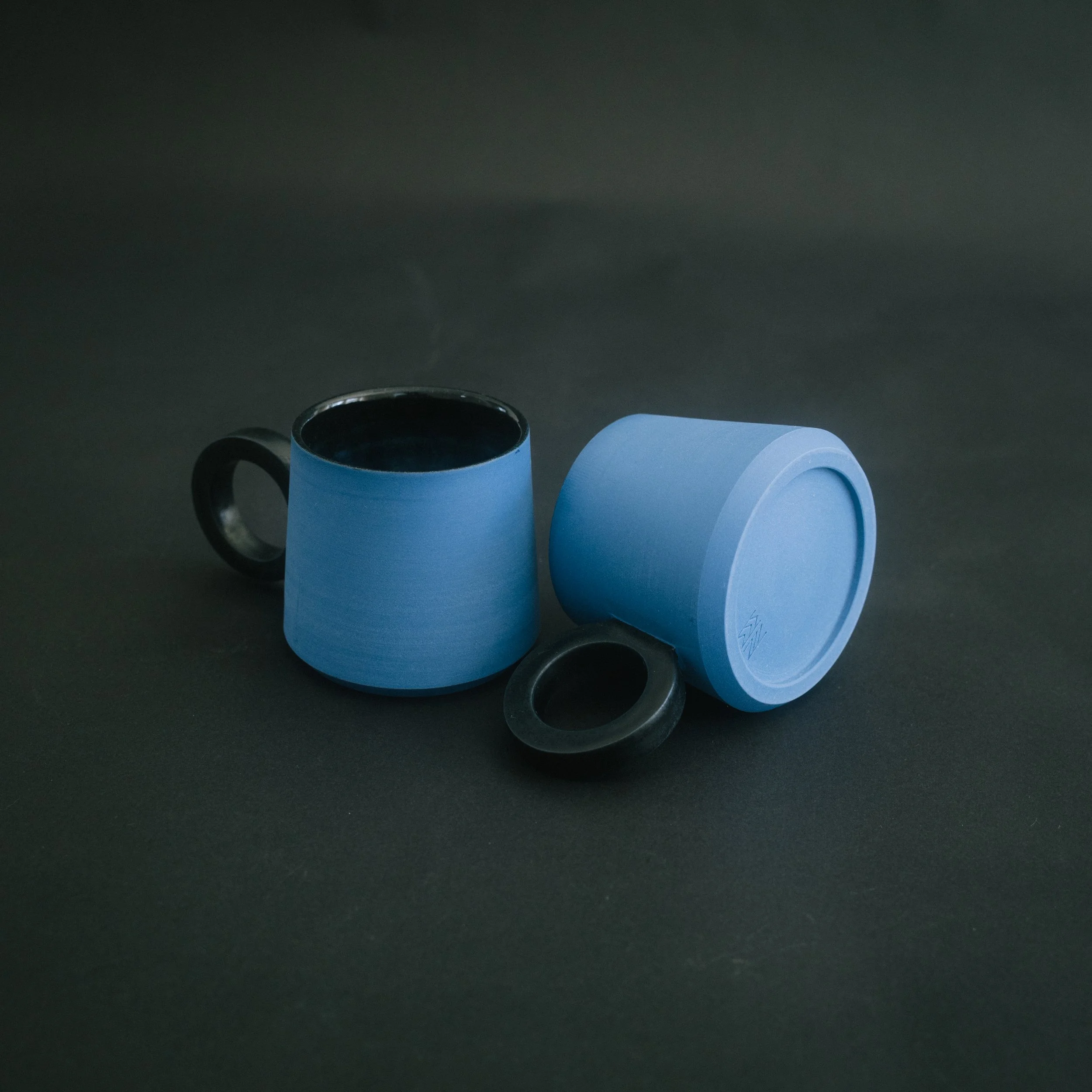 2 blue ceramic espresso cups with black handles on a black seamless background.