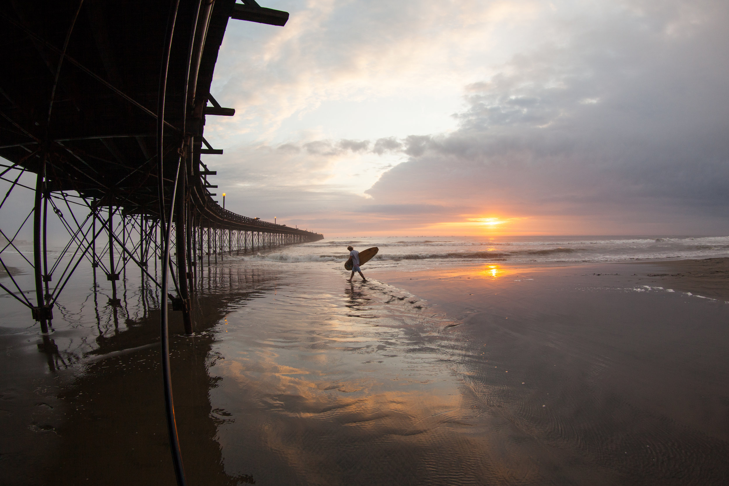 A surfer walks along the beach in Chiclayo with it's interesting curved pier whilst the sun sets over the ocean behind.