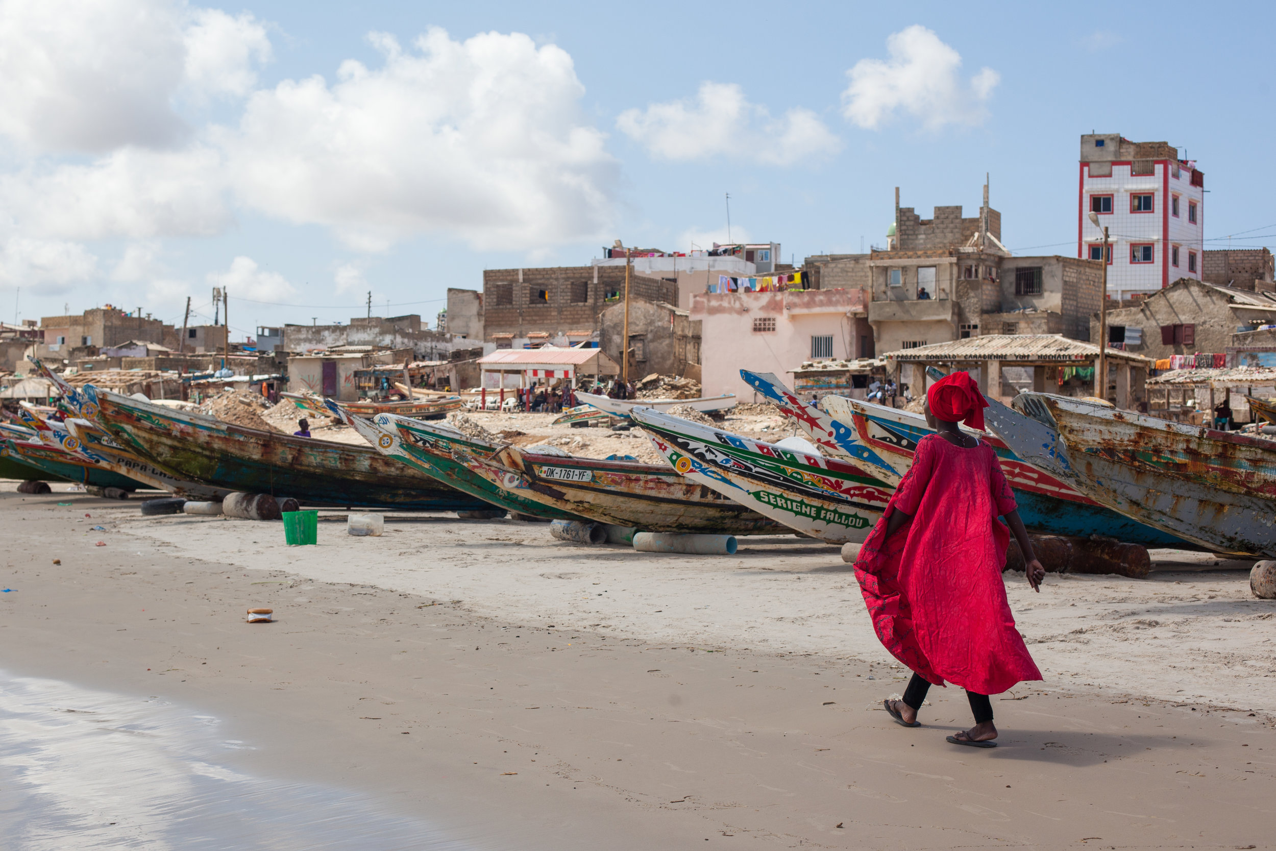 A Sengalese woman in striking African clothing walks past the fishing boats which line the beach.