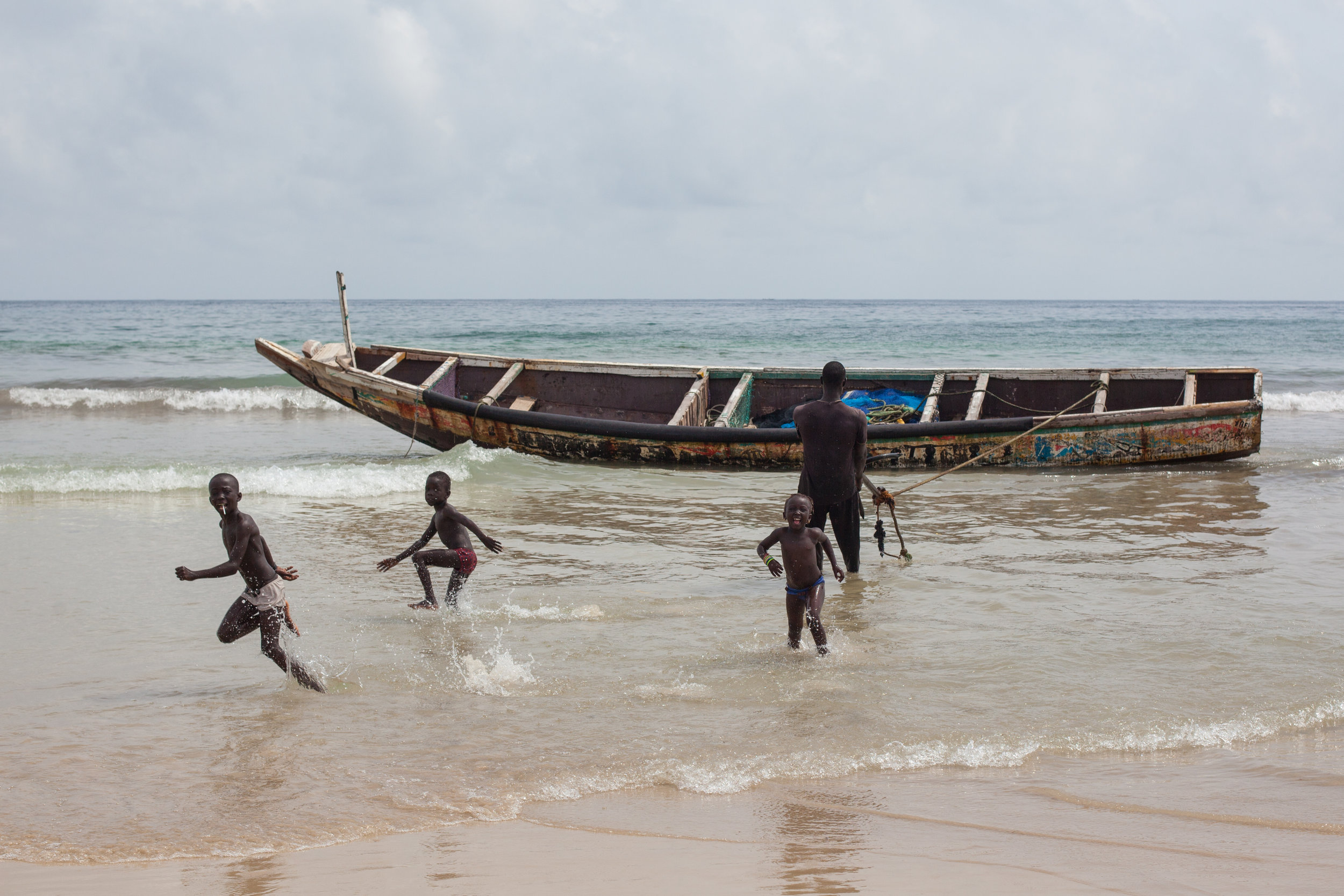 Excited African children play in the ocean as their father brings his boat back onto shore.