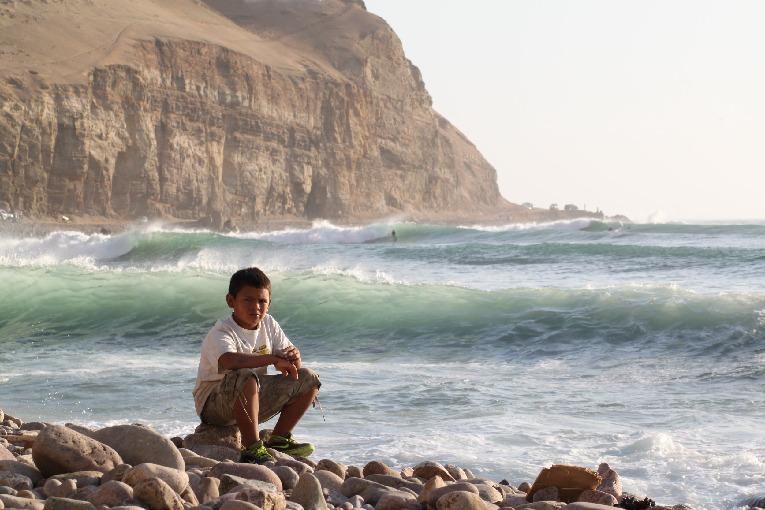 A boy sits on the shore of La Herradura, Lima, while waves are surfed in the distance.