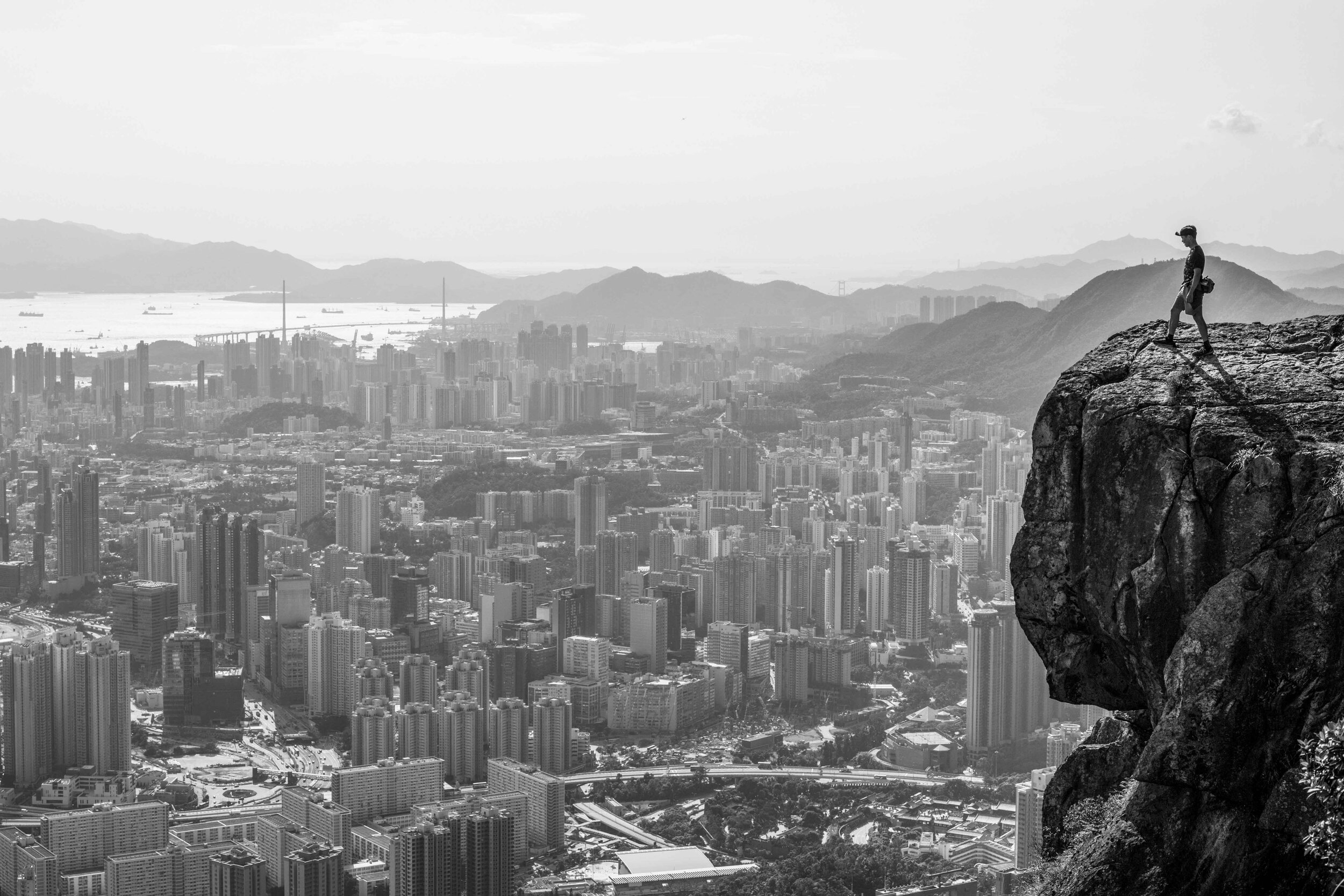 Black and white travel image of a hiker on a rock face high above Hong Kong.