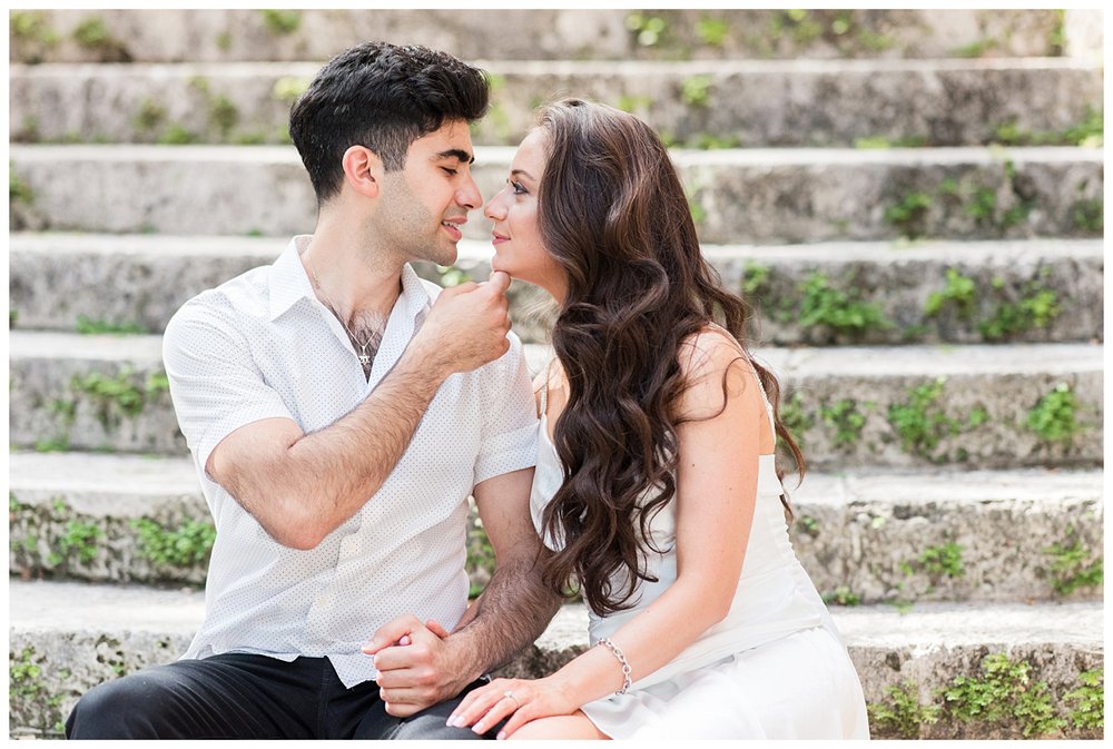 engaged couple in all white with groom's hand on bride's chin sitting on stairs