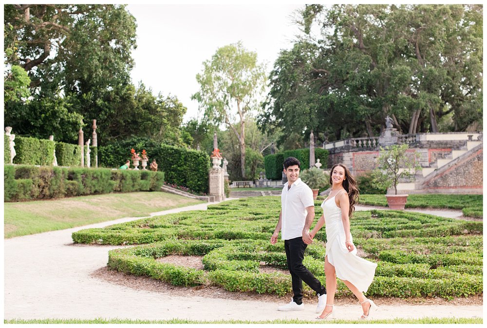engaged couple in white dress and white shirt walking through the gardens at Vizcaya Museum in Miami, Florida