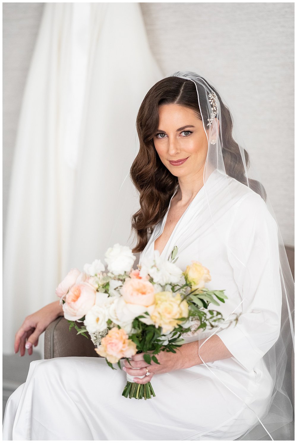 bride in white robe posing for Boston wedding photographer sitting in front of wedding gown holding floral bouquet