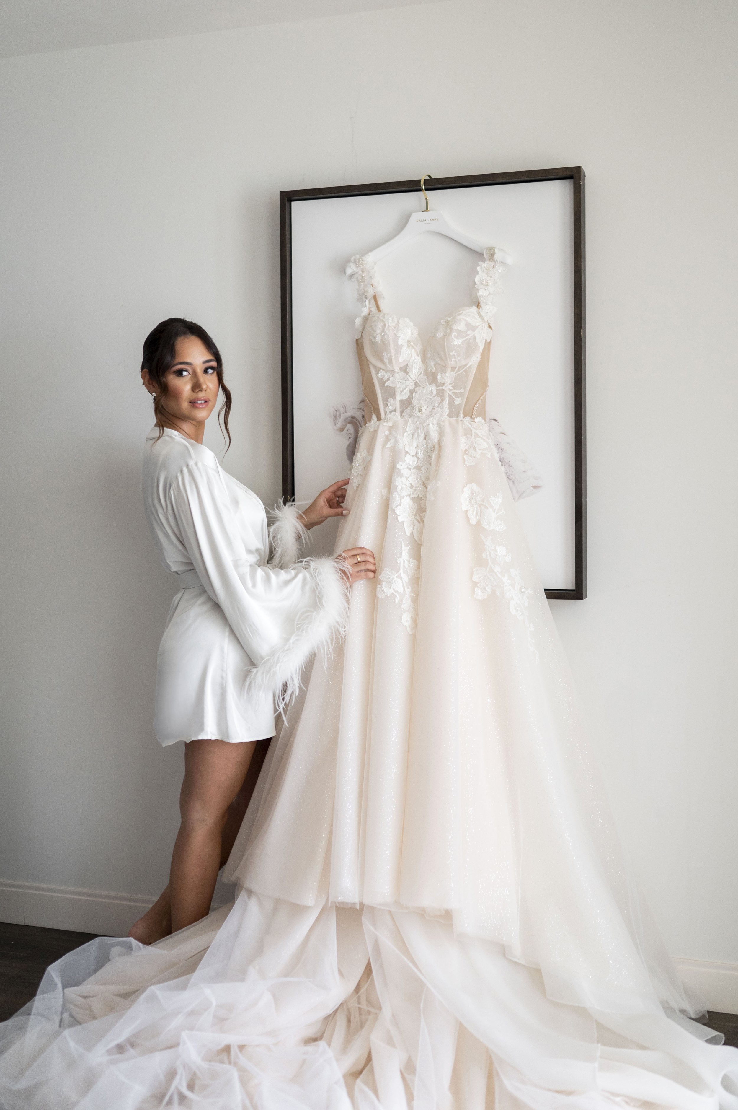 bride posing  next to her gown hanged on the wall