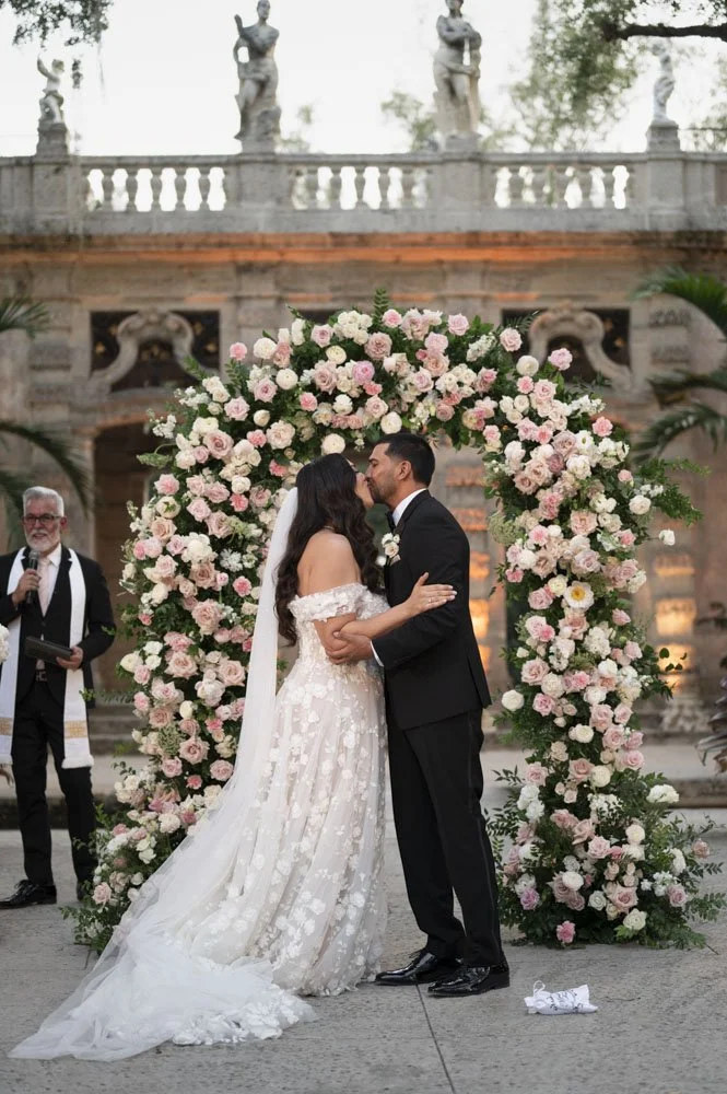 bride and groom kissing as they are declaire husband and bride in front of ther flower arch at the vizcaya museum and gardens in miami