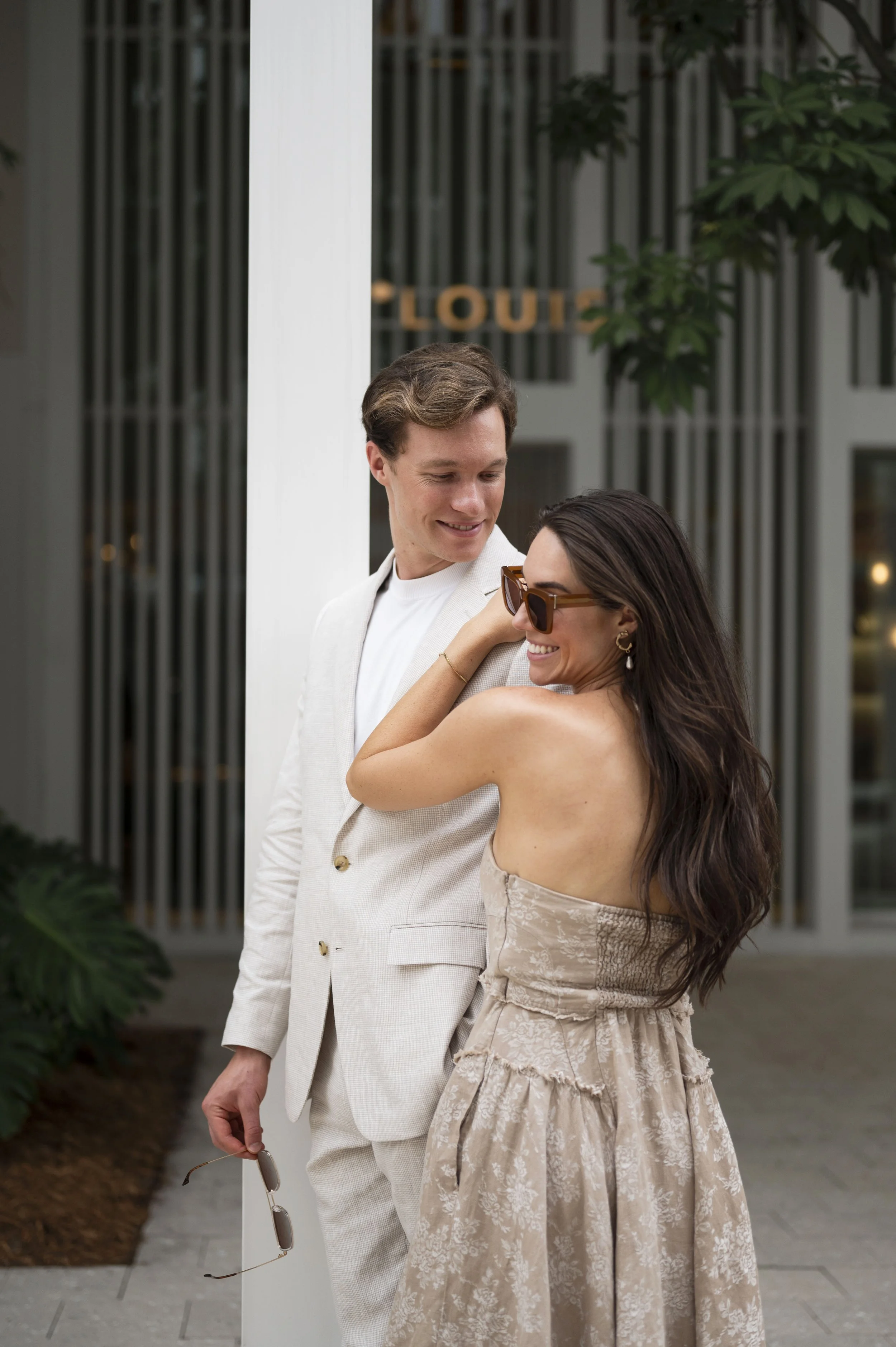 Travis looking at Rachel as she laughs wearing sun glasses both leaning to the Hermes store in the miami design district during their engagement session