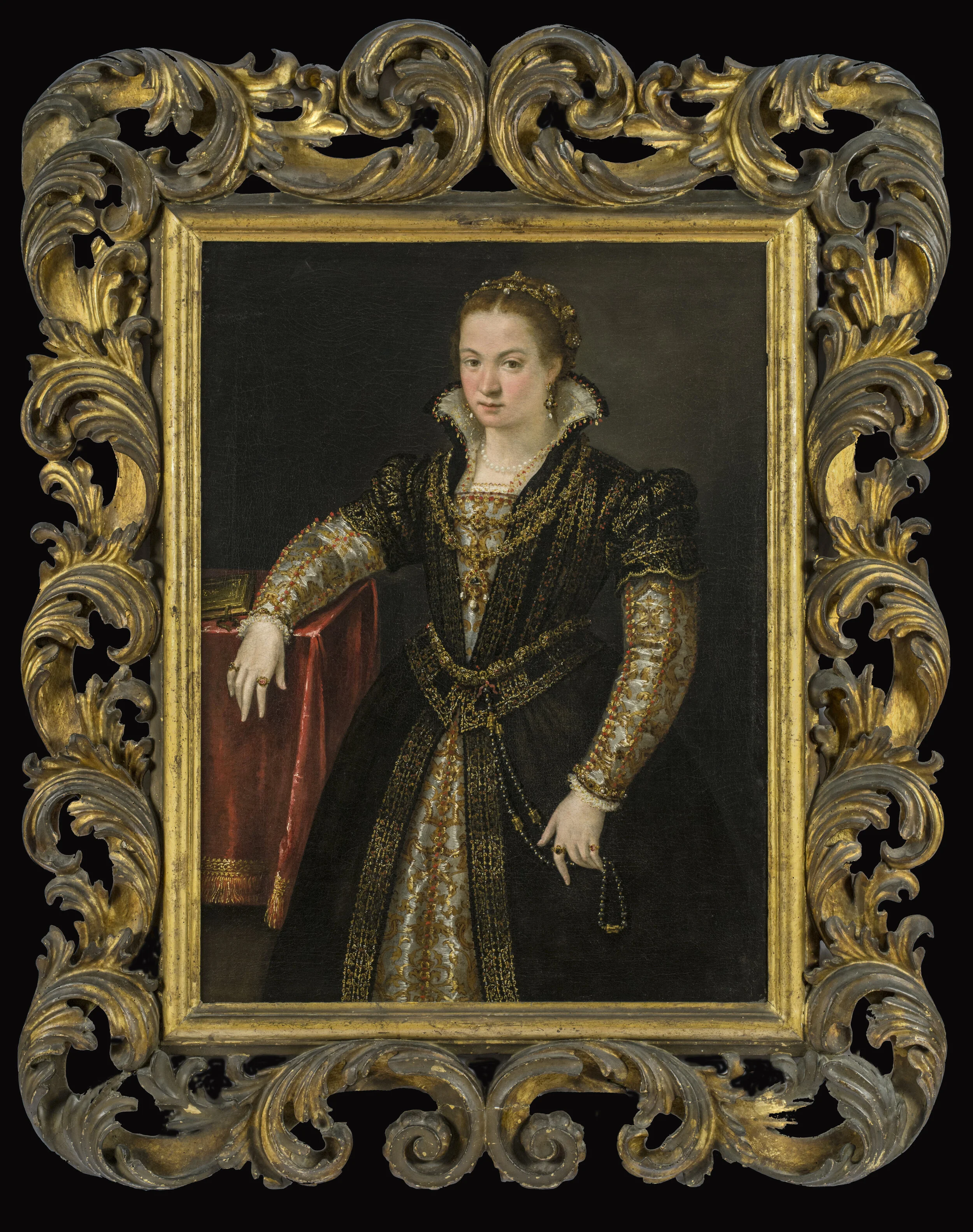 A noblewoman in a brocade and silk dress rests her right elbow on a table with a red cloth.