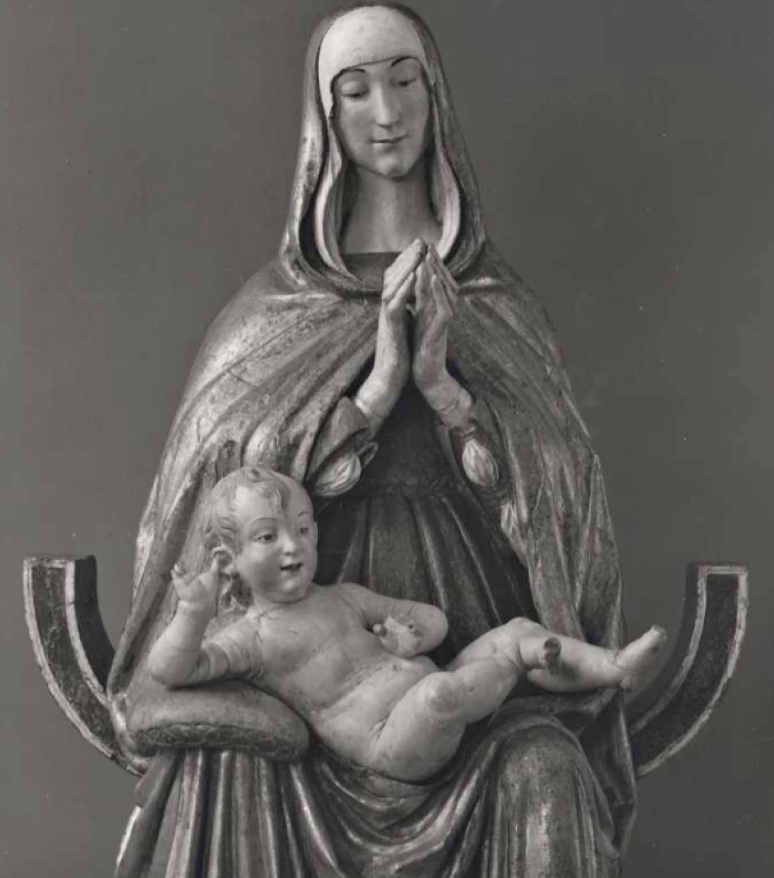 Woman wearing a robe with her hands folded in prayer, with a child reclining in her lap.