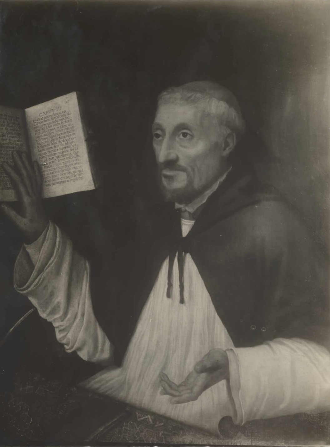 Black-and-white image of a man sitting at his desk covered with a carpet while pointing at a book and giving a lecture.