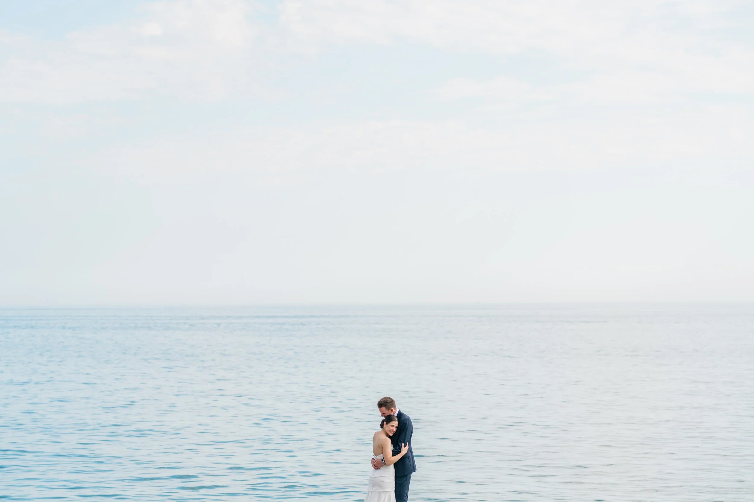 A couple in wedding attire embracing by the water on a cloudy day.