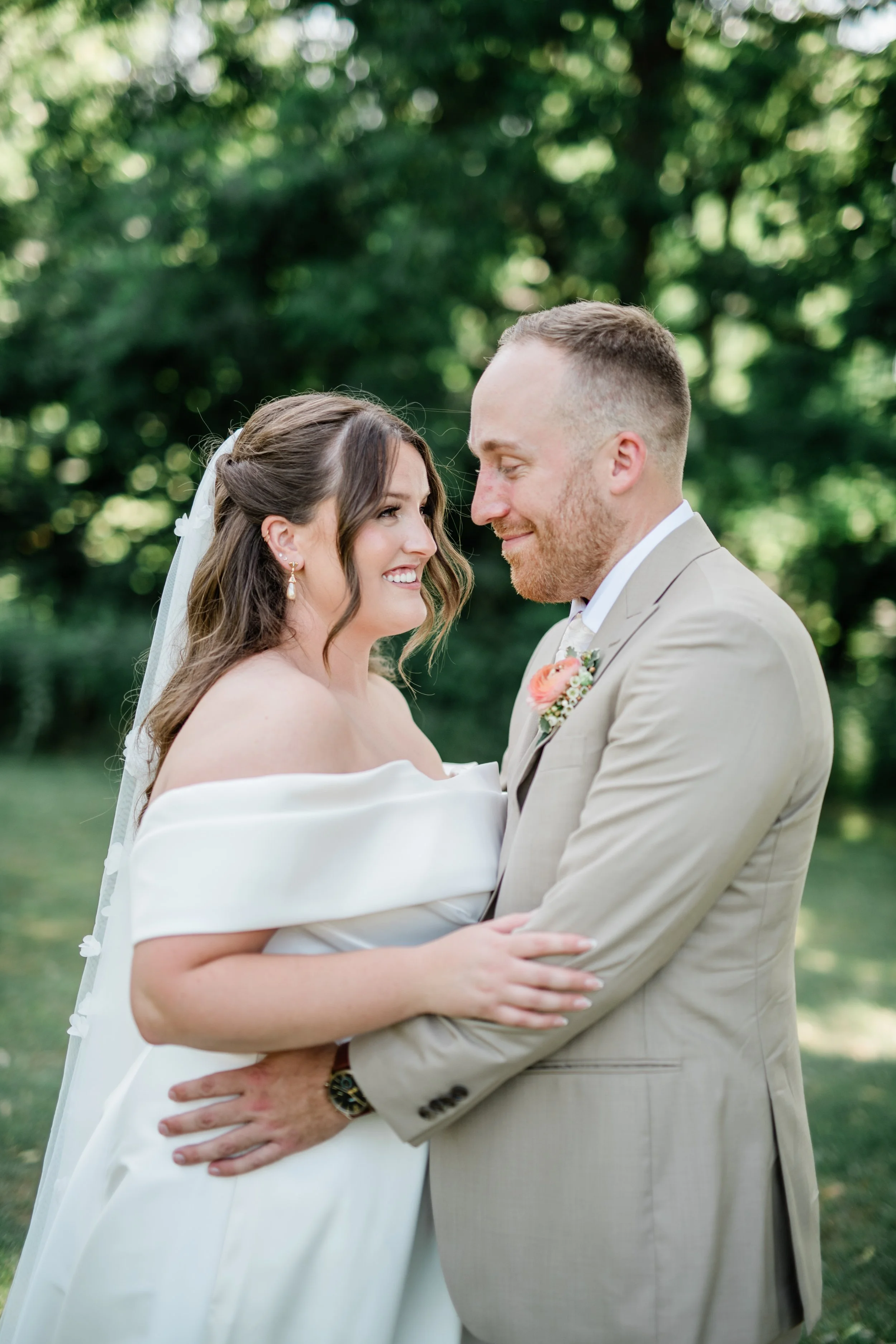 A bride and groom stand close together outdoors, smiling and gazing into each other's eyes, with trees in the background.