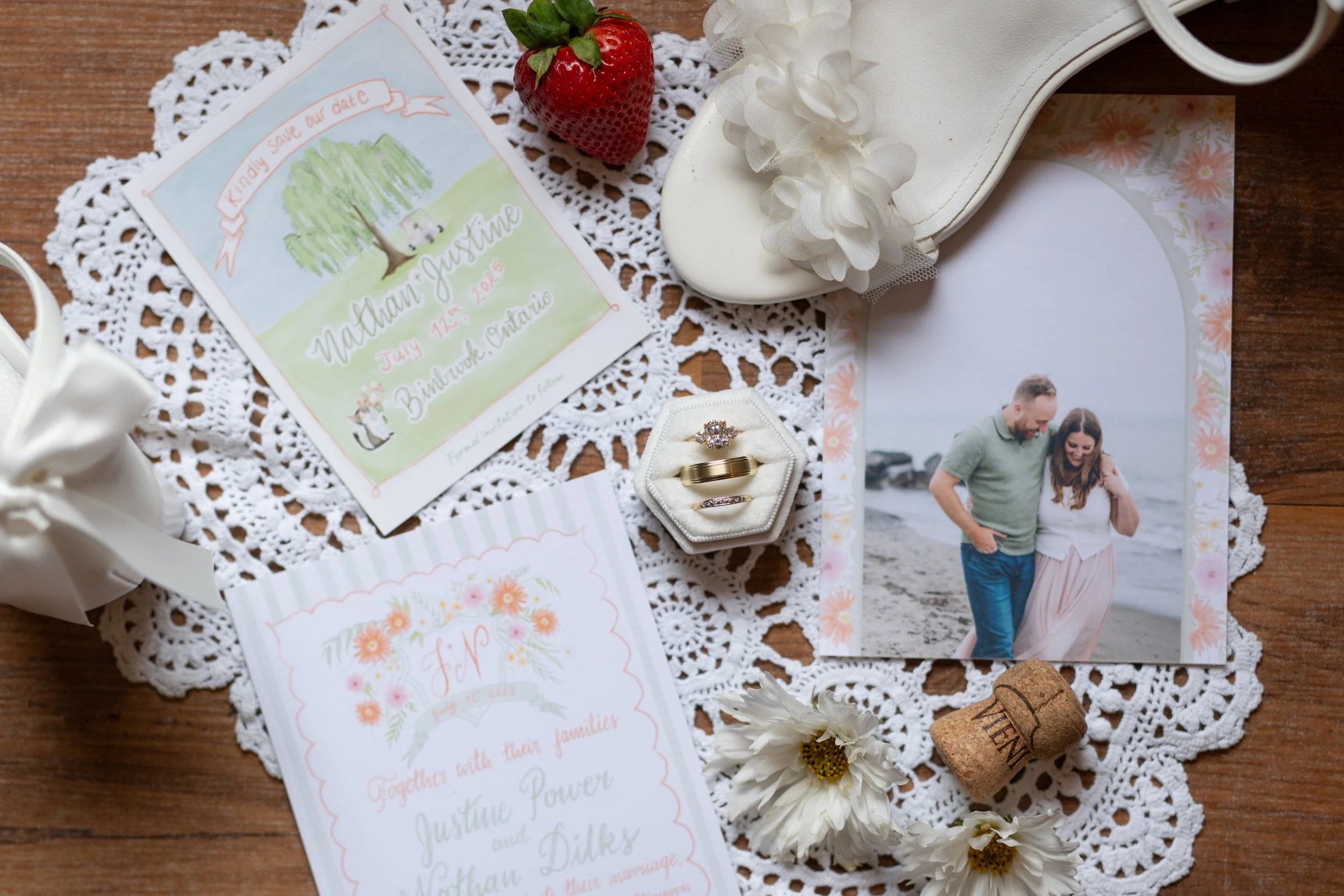 Wedding flat lay with invitation, photograph of a couple, wedding bands in a ring box, strawberries, white flowers, a cork, and lace and paper decorations on a wooden surface.