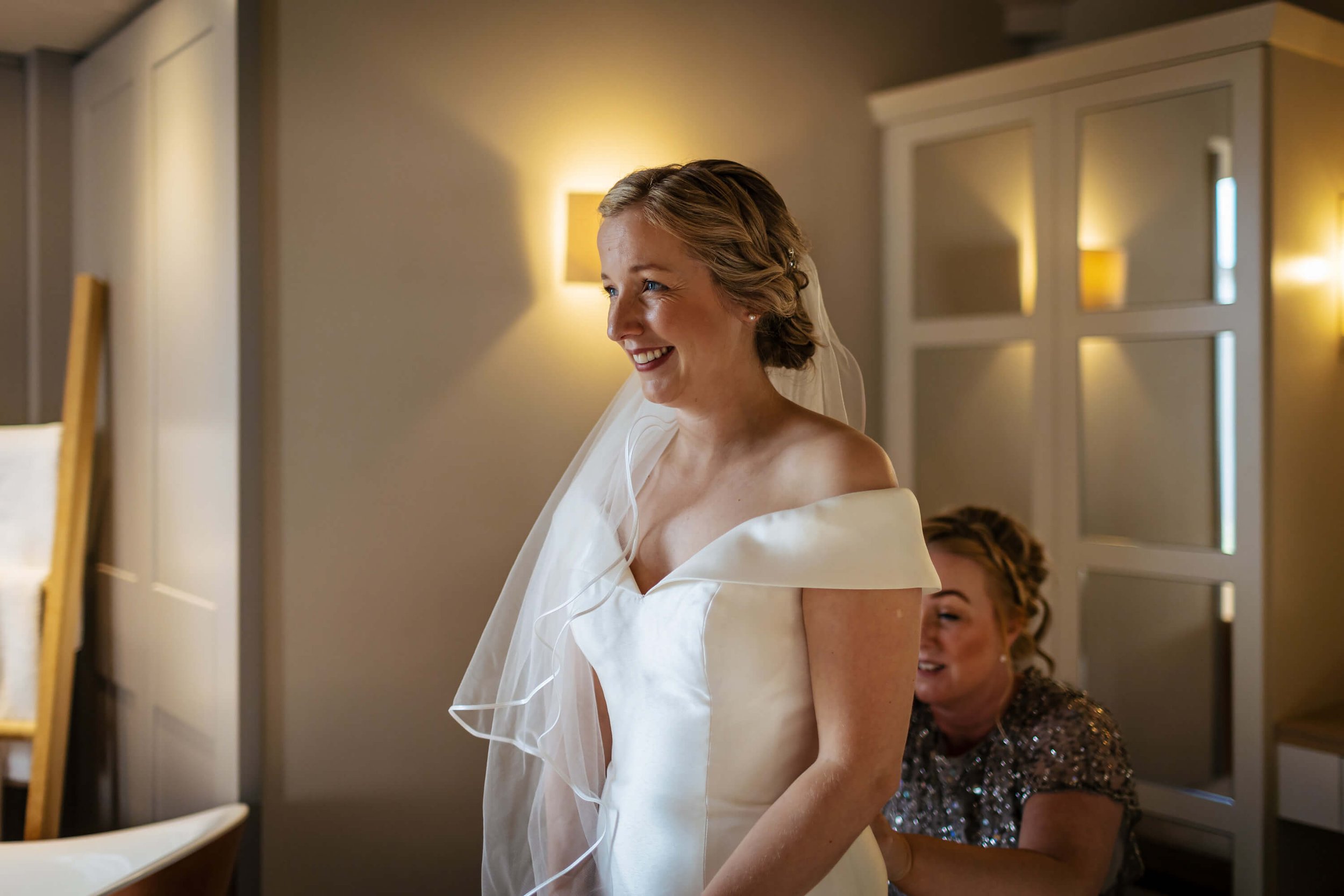 Bride smiling while the bridesmaid helps her dress up at the back