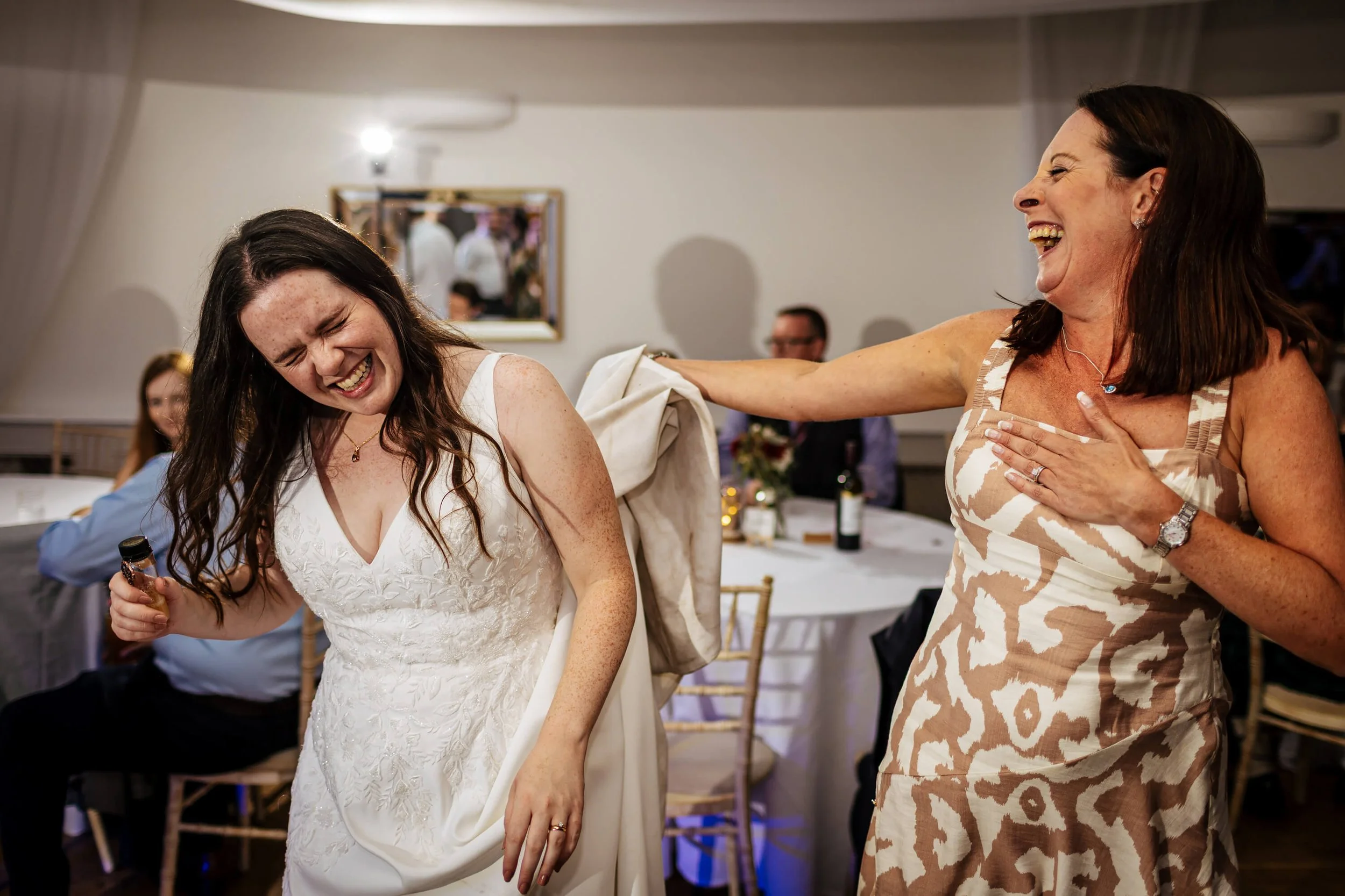 Bride laughing on the dance floor as a guest holds up her dress to help